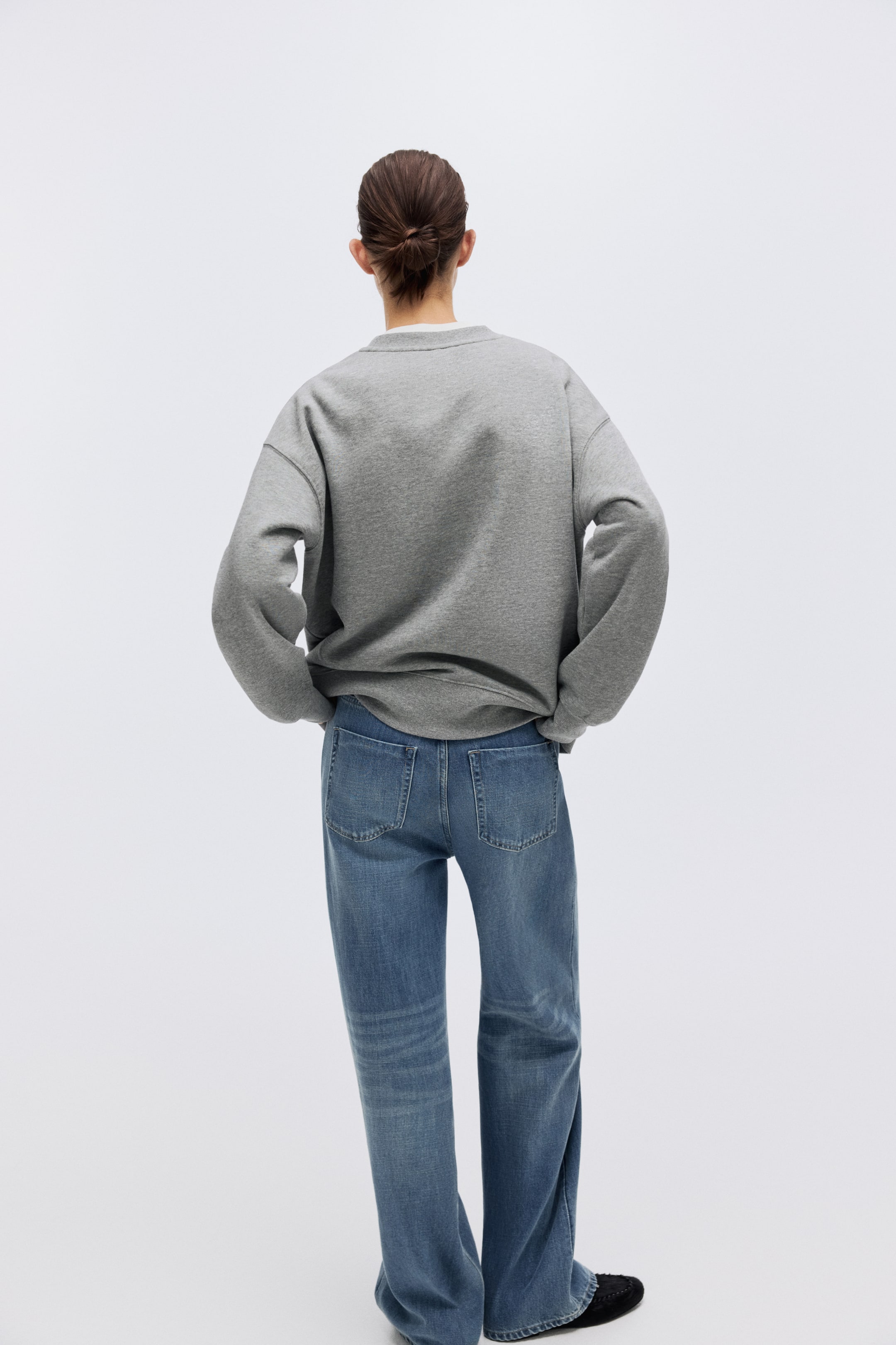 View larger image: A grey crew-neck sweatshirt is shown from the back, featuring an oversized fit with voluminous sleeves, and its ribbed hem gathers at the waist. It is paired with medium-blue, relaxed-fit jeans that show visible back pockets and subtle fading.