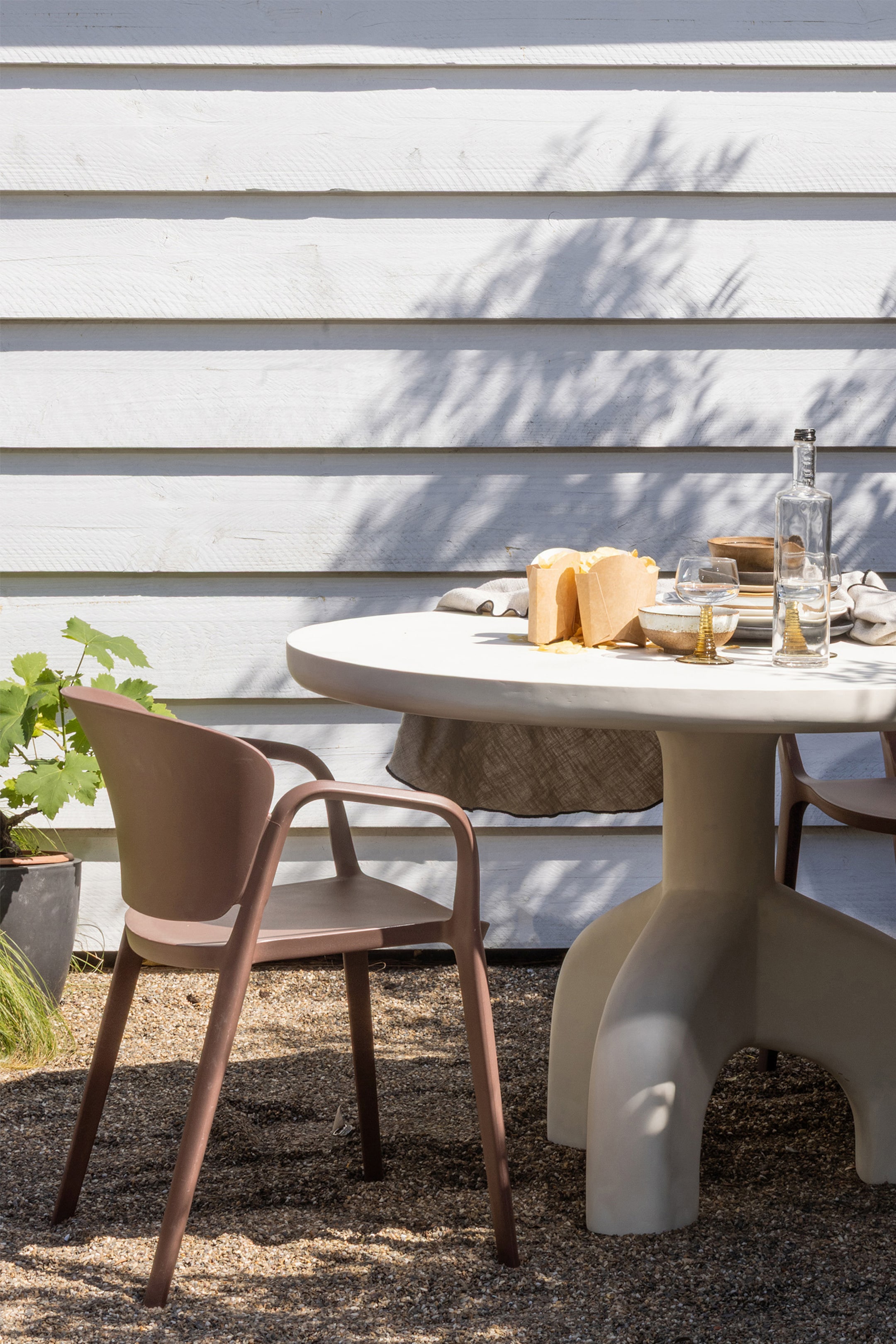 Agrandir l'image: Une table à manger ronde pour l'extérieur, avec un plateau de couleur crème et un pied central sculptural à trois pieds arrondis, est exposée. Deux chaises moulées marron, avec des dossiers incurvés et des accoudoirs intégrés, sont disposées autour de la table, qui est dressée avec des assiettes et des verres.