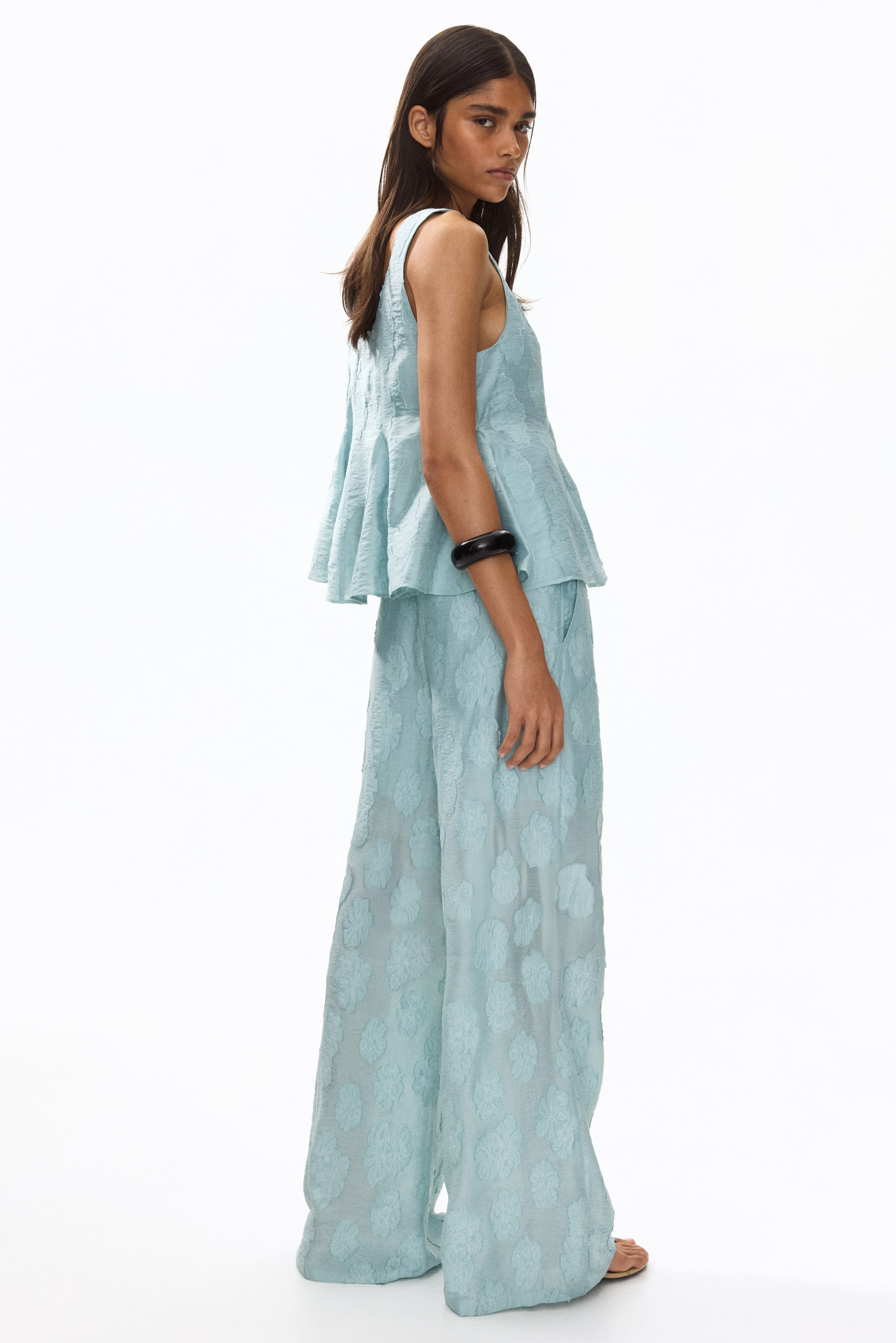 View larger image: The model is showcasing fitted light blue trousers, adorned with a floral jacquard weave, diagonal side pockets, and flared legs. A coordinating top is layered over the waist, while sandals peek out from beneath the flared legs.