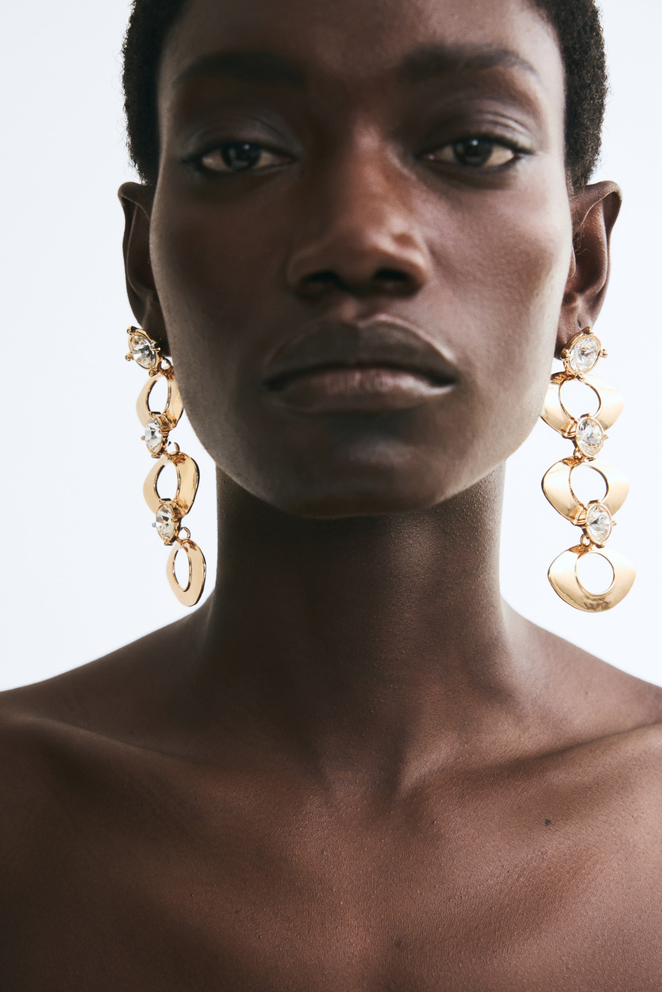 View larger image: Gold-toned metal dangle earrings featuring sparkling glass rhinestones and tiered, oval-shaped pendants with cut-out details are being worn by the model. The long statement earrings hang down on either side of the model's face, reaching past the jawline.