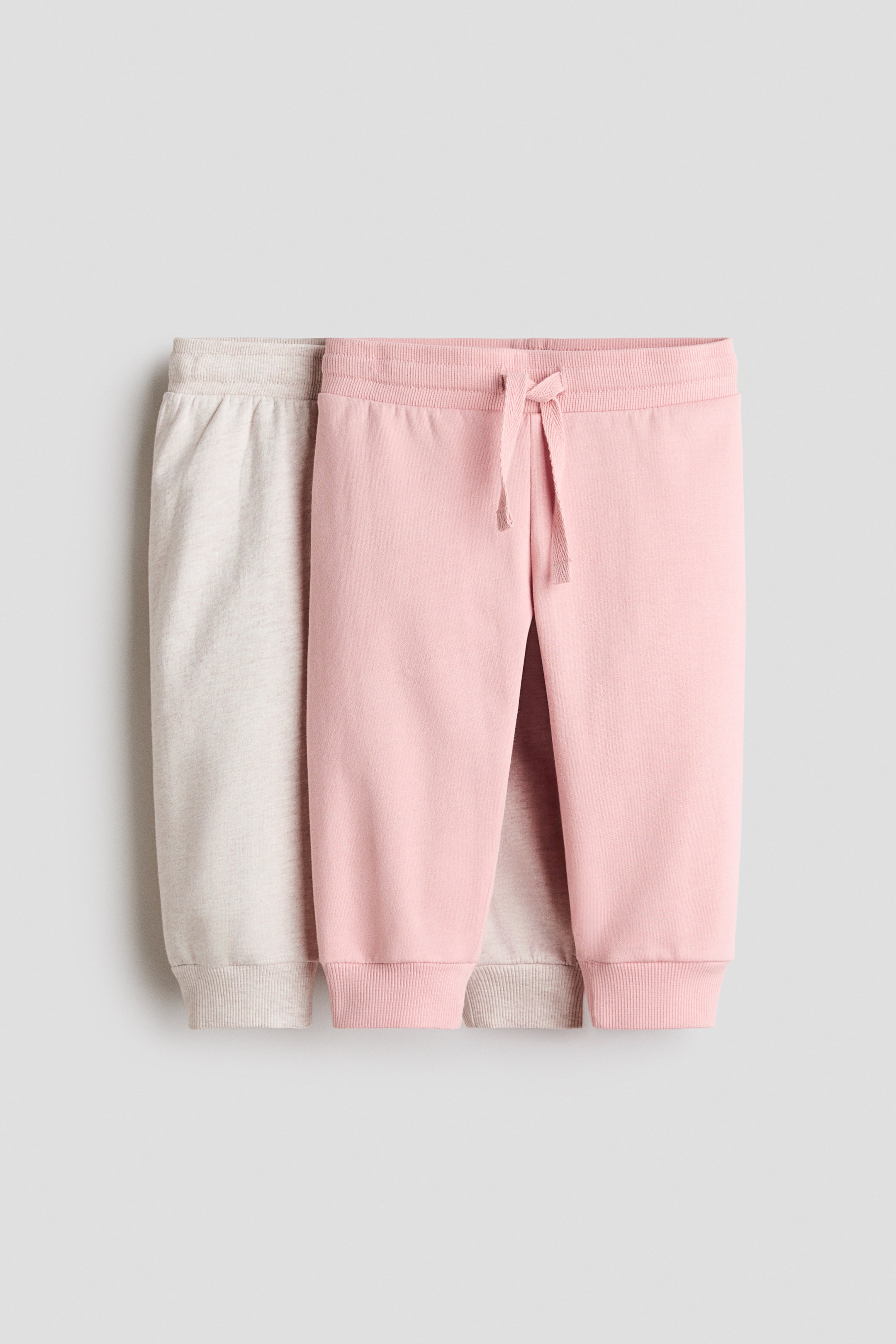 View larger image: "Dalawang pares ng sweatpants ang nakadisplay mula sa harap, isang light beige na pares na bahagyang nakikita sa likod ng pink na pares. Parehong pares ay may ribbed waistbands at ribbed ankle cuffs, at ang pink na pares ay may nakikitang tali sa harap ng baywang."