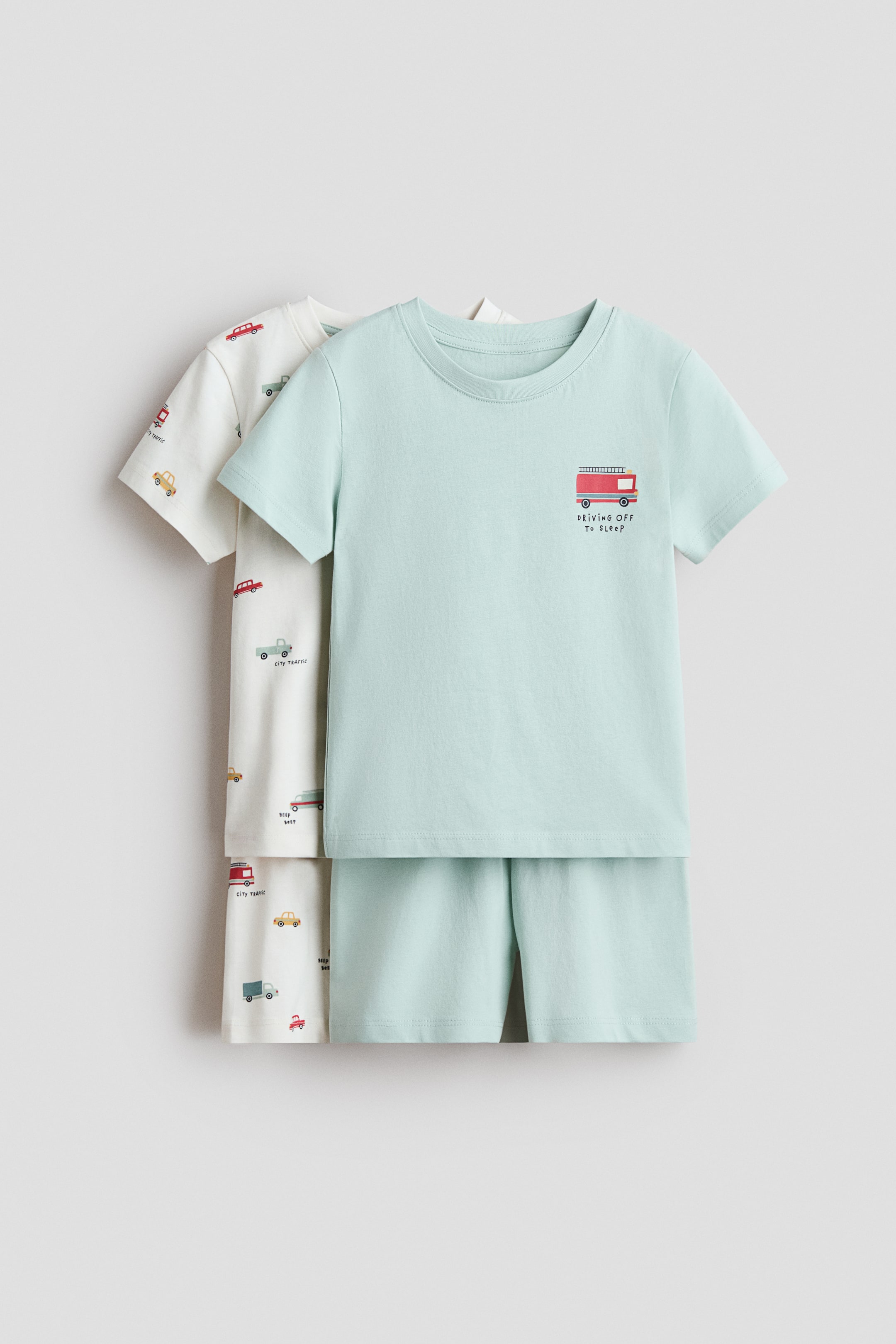 View larger image: A set of pyjamas, facing front, showcases a light blue T-shirt with a ribbed neckline and a small fire truck graphic on the front, paired with matching shorts featuring a covered elasticated waist. A second cream pyjama set, adorned with an all-over vehicle print, is partially visible in the background.