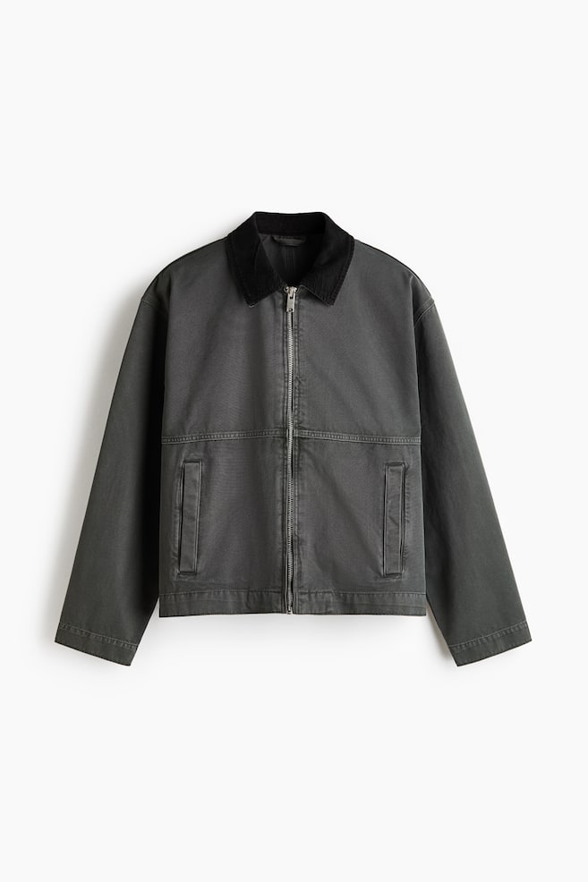 Une veste de travail en toile de coton gris foncé est présentée de face, avec un col à revers en velours côtelé noir contrastant. Une fermeture éclair argentée descend au centre, avec deux poches passepoilées rectangulaires visibles sur le bas, et des manches longues s'étendent jusqu'à un ourlet droit. - 1