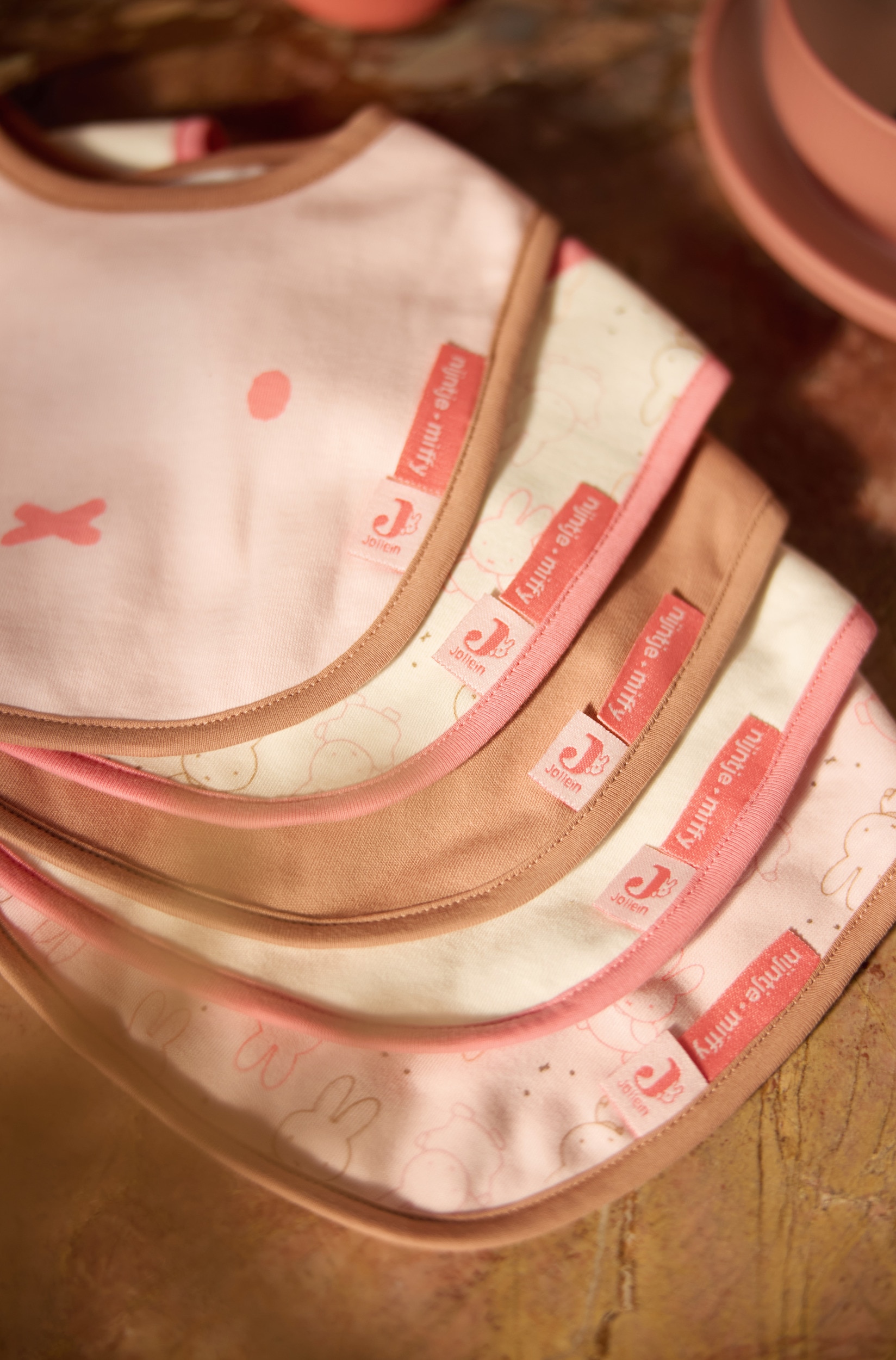Vijf slabbetjes voor baby's liggen overlappend in een diagonale stapel en tonen een kleurenpalet van lichtroze, crÃ¨me en bruin. Het voorste slabbetje heeft een effen lichtroze stof met een vereenvoudigde konijnenprint en bruine afwerking, terwijl de slabbetjes erachter bijpassende patronen, effen kleuren en klittenbandsluitingen aan de hals laten zien.