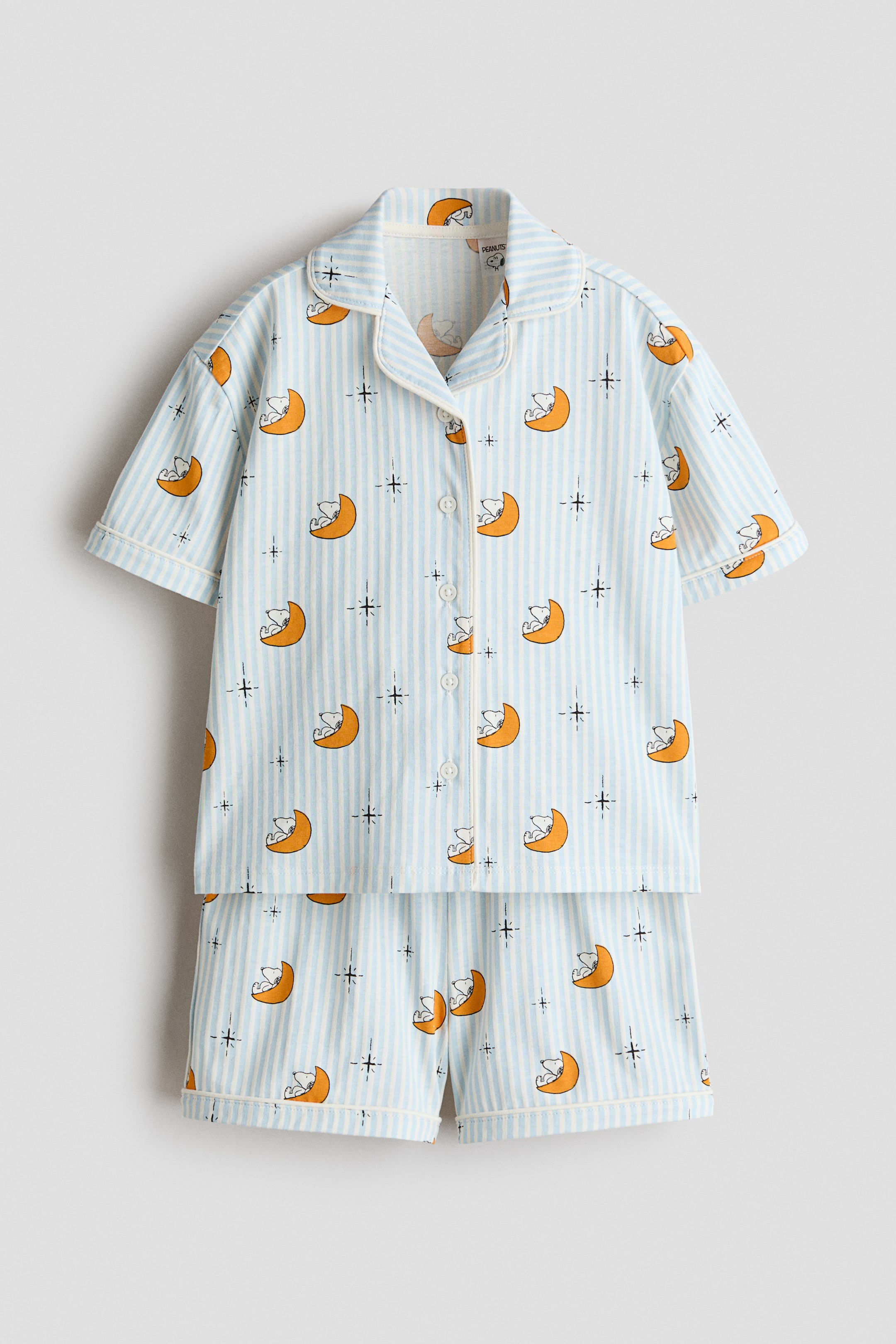 View larger image: This pajama set in light blue and white stripes is showcased front-on, adorned with a charming pattern of Snoopy dozing on orange crescent moons alongside delicate starbursts. The short-sleeved top features a notch lapel collar and a button-front design, while the coordinating shorts complete the ensemble. Light yellow piping elegantly outlines the collar, front placket, sleeve cuffs, and hem of the shorts, adding a refined touch.