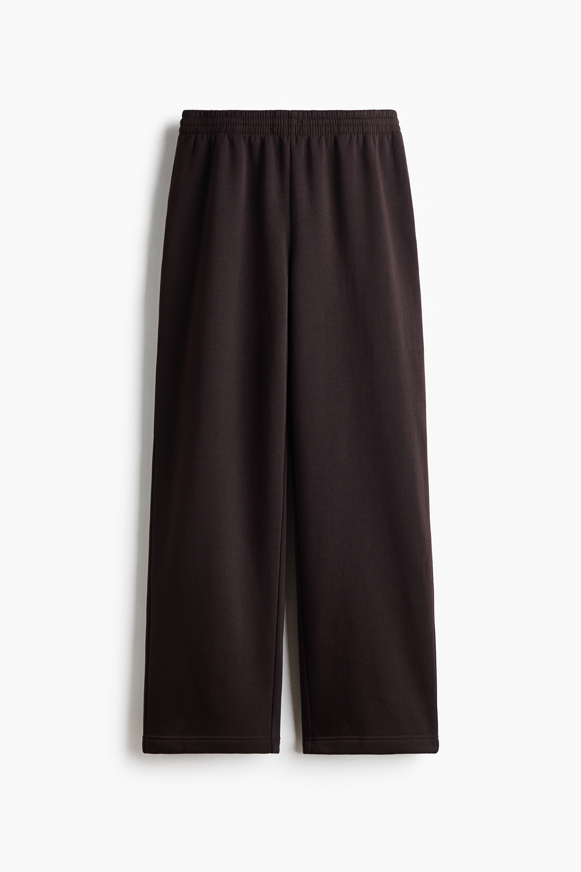 Track pants in a dark brown sweat fabric are being worn by the model, featuring straight legs and an elasticised waistband with an adjustable drawstring. The pants are styled with a matching dark brown zip-up hoodie, a white top, and black sandals.