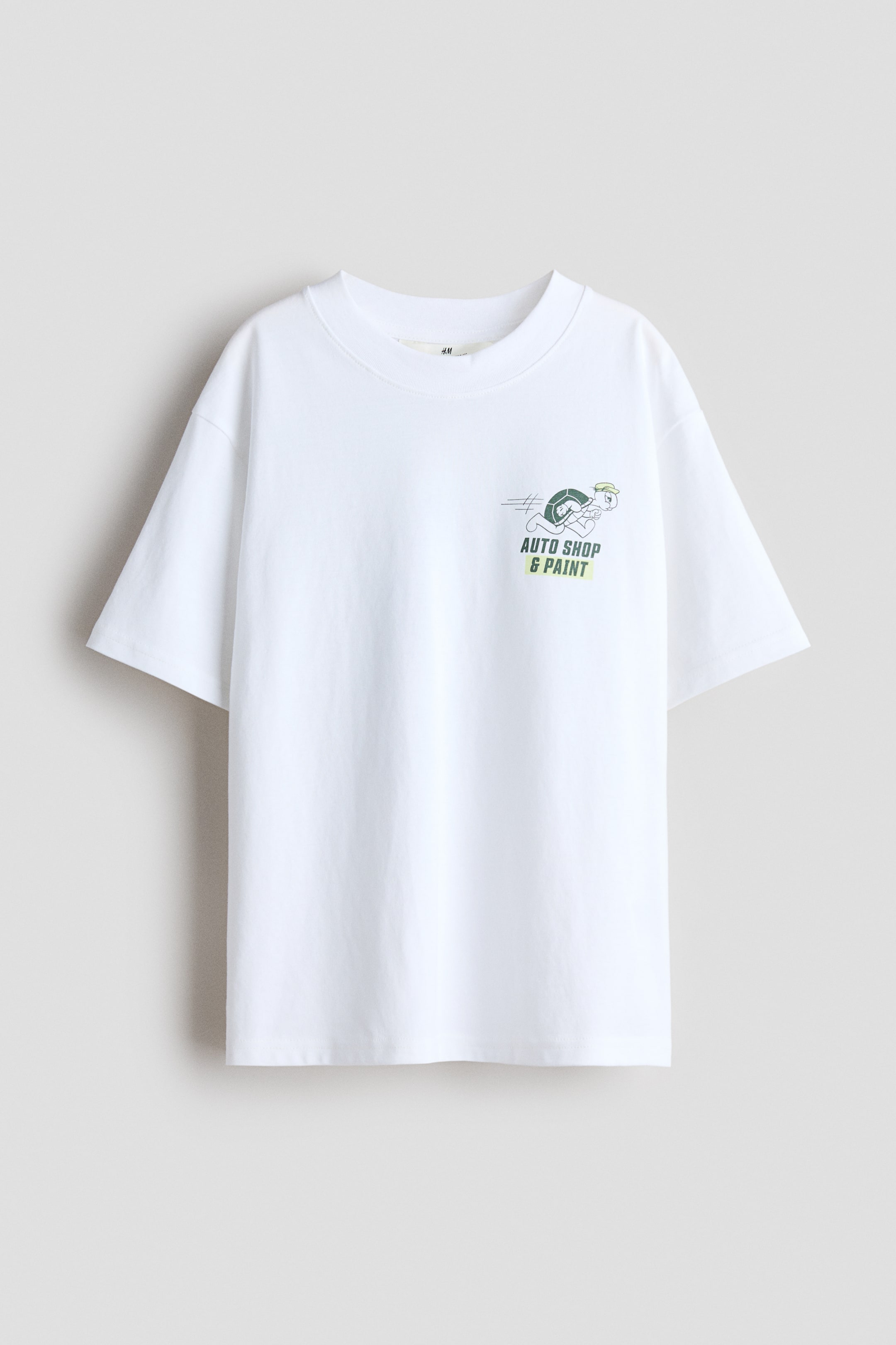 Größeres Bild ansehen: Die Vorderseite eines weißen, locker sitzenden T-Shirts aus bedrucktem Baumwolljersey ist sichtbar, mit einem gerippten Besatz am Halsausschnitt und überschnittenen Schultern. Ein grüner, hellgrüner und schwarzer Grafikdruck, der eine Cartoon-Schildkröte zeigt, die einen Autoreifen mit dem Text "auto shop & paint" darunter schiebt, erscheint auf der linken Brust.