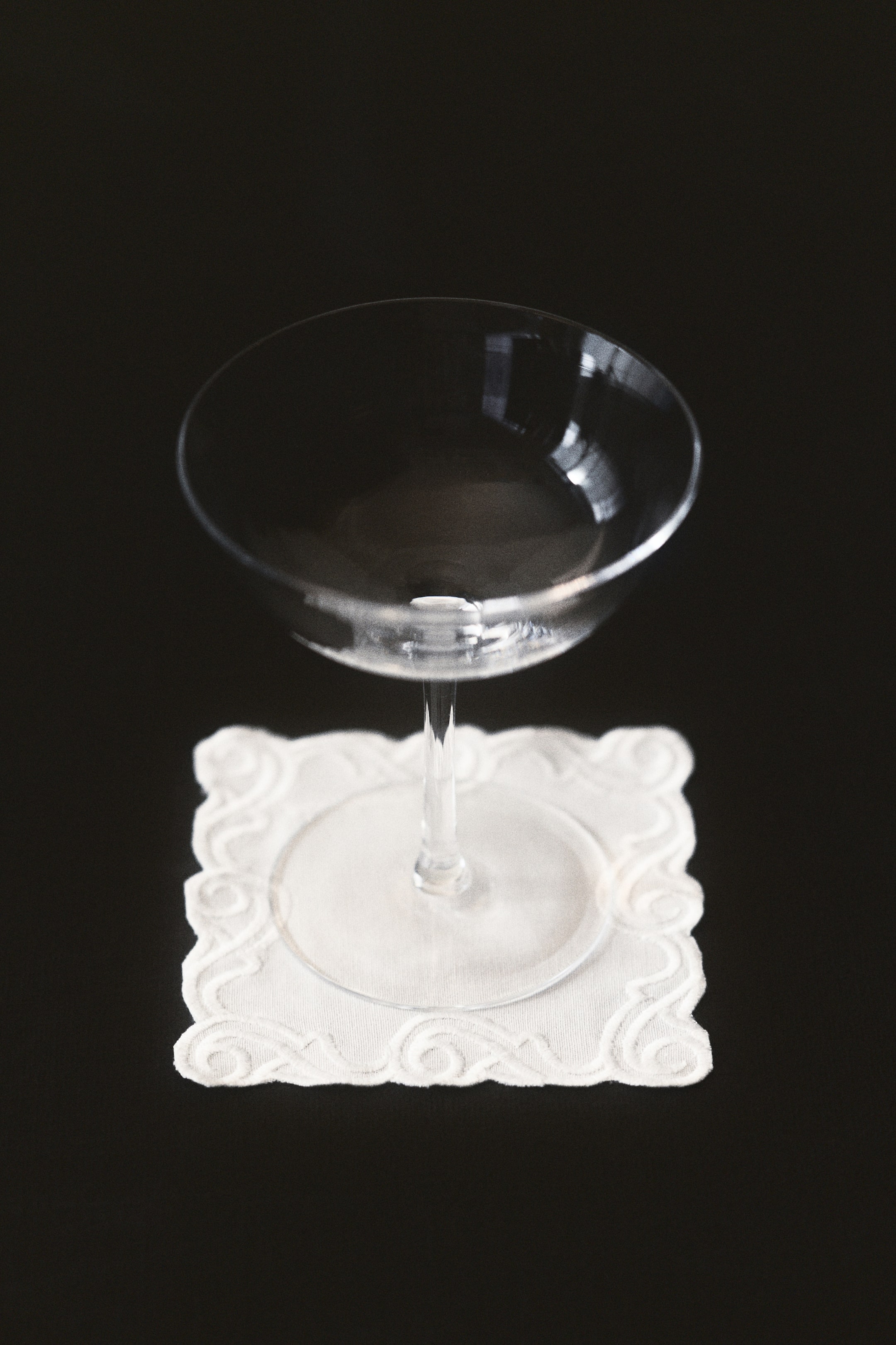 View larger image: A square coaster showcases a cotton weave texture in a light cream colour. It is detailed with a tone-on-tone embroidered swirling pattern along its scalloped edges.