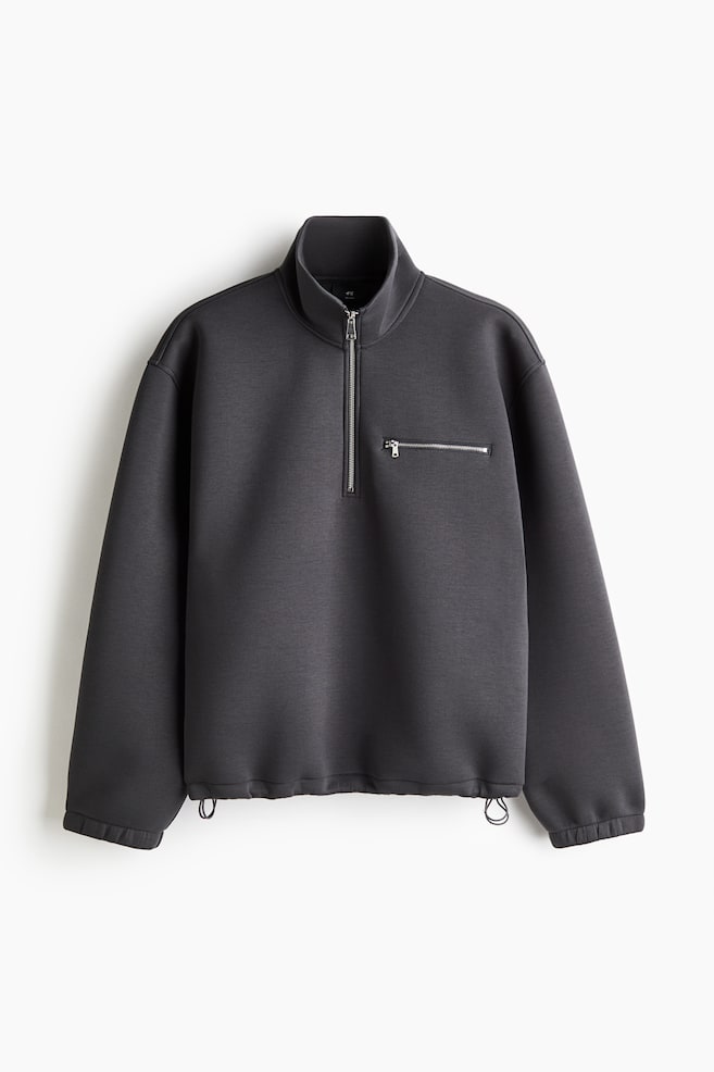 En mÃ¸rkegrÃ¥ sweater i scuba-stof vises forfra og har en opretstÃ¥ende krave med halv lynlÃ¥slukning samt en vandret brystlomme med lynlÃ¥s. Plagget har sÃ¦nkede skuldre, lange Ã¦rmer med elastiske manchetter og en justerbar kant med snÃ¸re og synlige snorestoppere. - 1