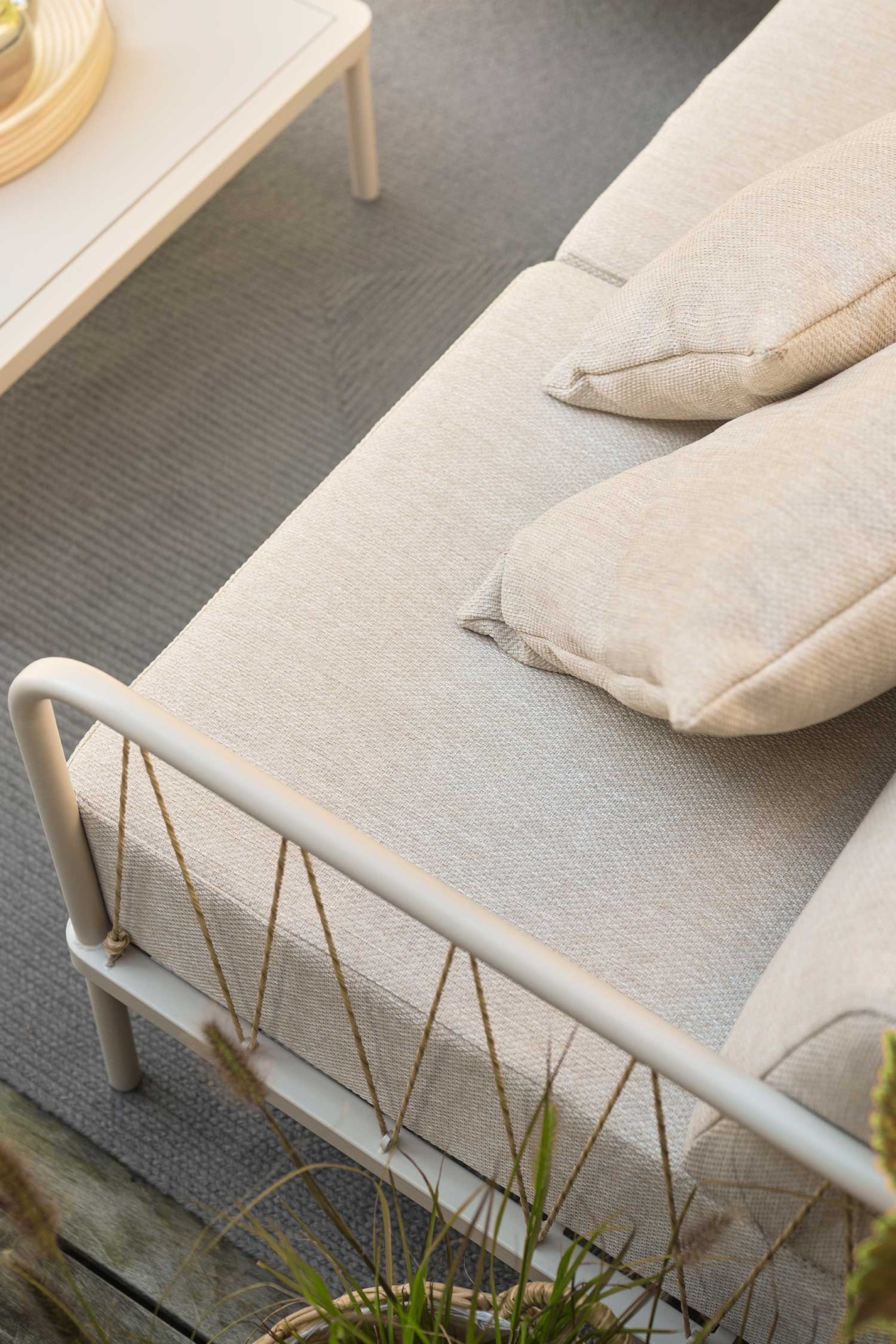 Größeres Bild anzeigen: Ein Outdoor-Sofa mit einem Sitzpolster aus hellbeigem, gewebtem Stoff, auf dem zwei kleinere Rückenkissen ruhen. Der hellfarbige, röhrenförmige Metallrahmen zeigt ein Seitenpaneel, und diagonale Seile erstrecken sich zwischen den oberen und unteren Schienen.