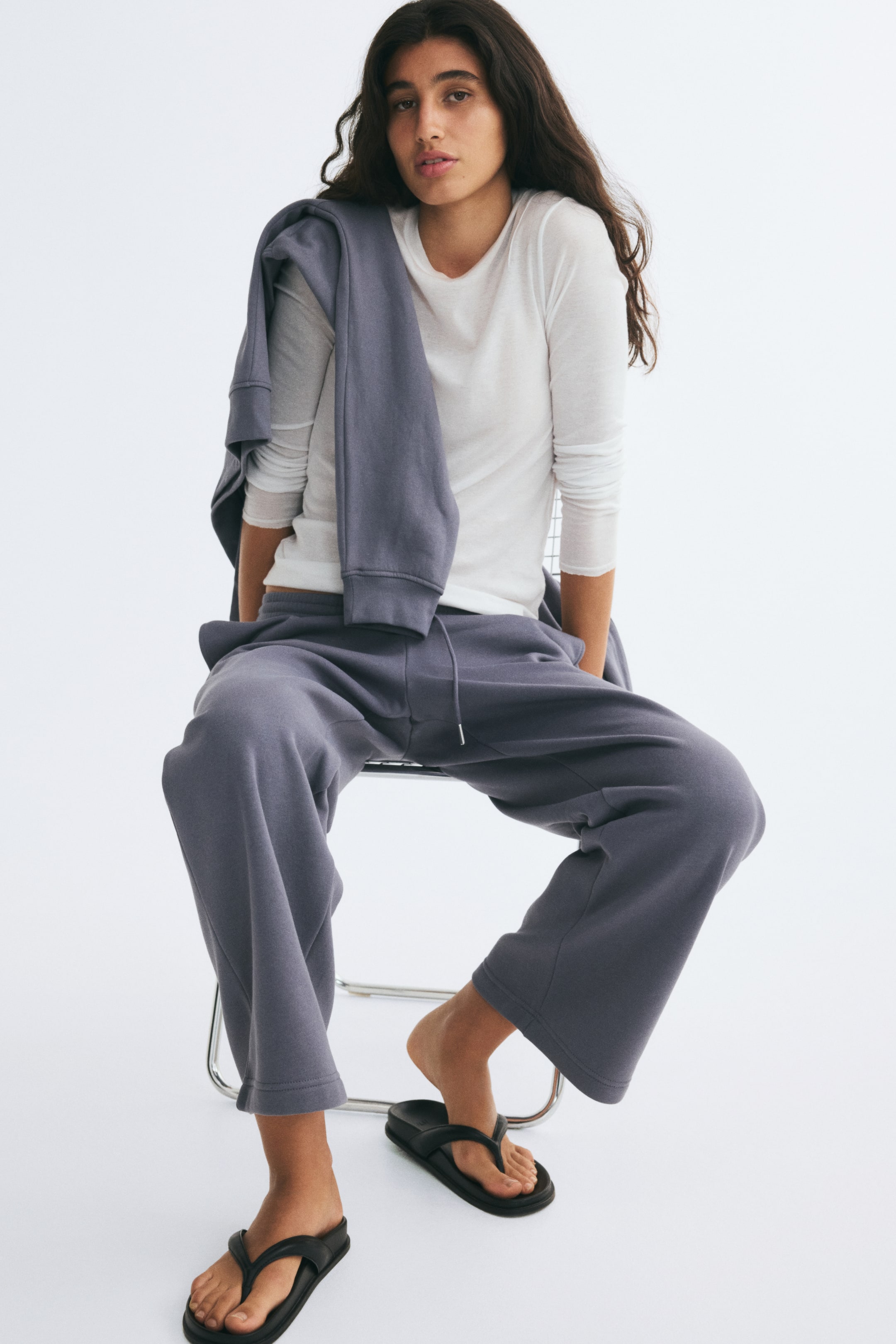 Ampliar la imagen: La modelo lleva un pantalón deportivo en tejido sudadera azul grisáceo apagado, con un corte recto relajado y cintura con cordón ajustable. El pantalón se combina con una blusa blanca de manga larga, una sudadera a juego colocada sobre el hombro de la modelo y sandalias planas negras.