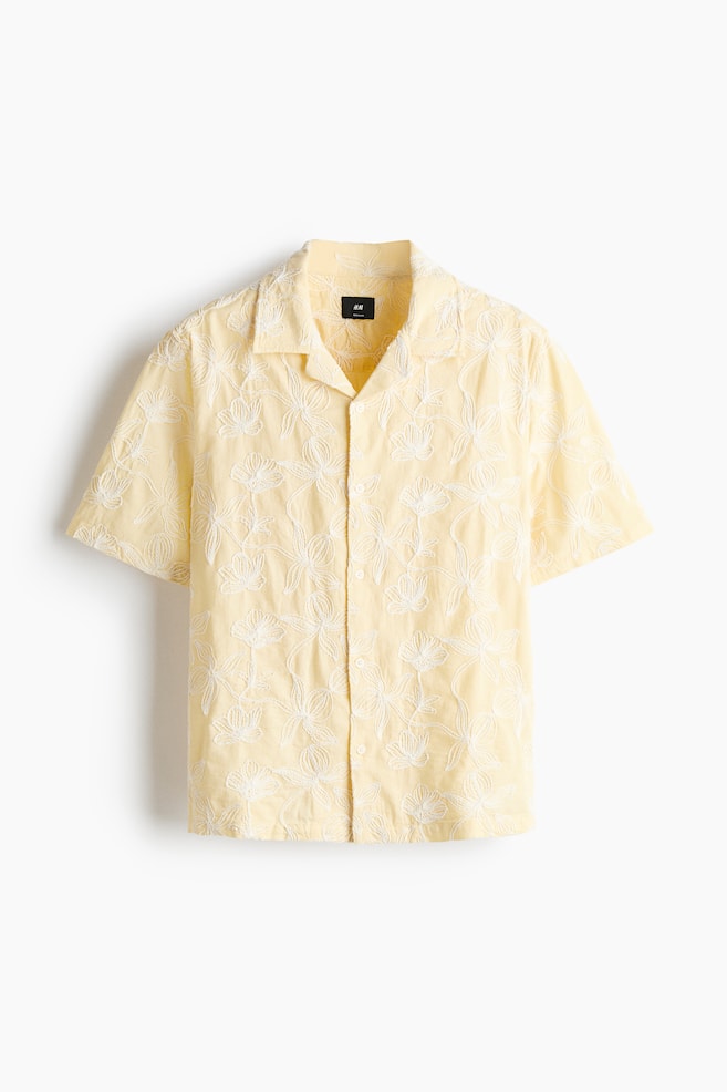 A yellow short-sleeved shirt is displayed front-facing, featuring a revere collar and short sleeves. White floral embroidery adorns its entire surface, with white buttons running down the front placket. - 1