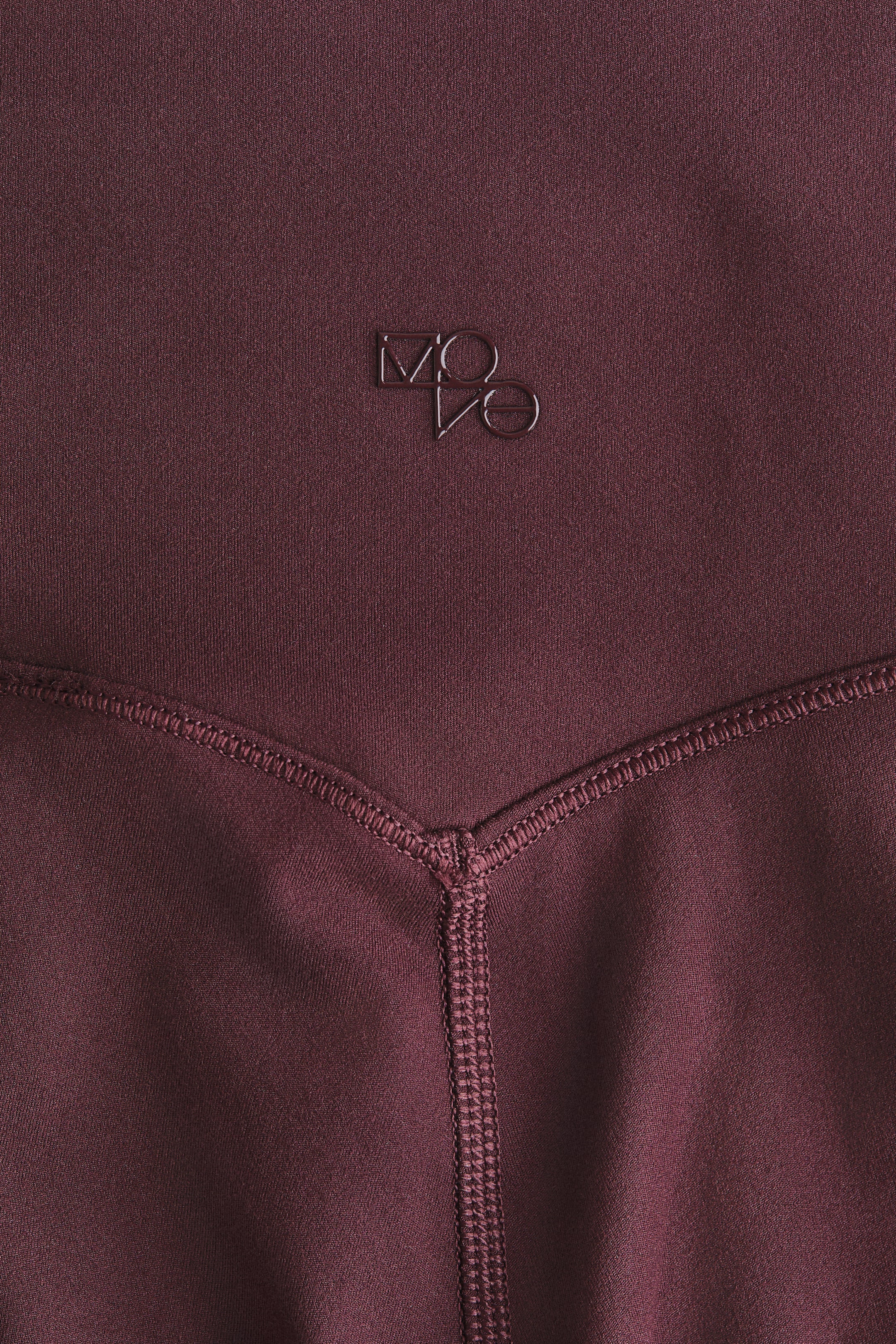View larger image: The front of the sports leggings is displaying its deep burgundy fabric. A metallic 'MOVO' logo is visible on the upper section, with a V-shaped flatlock seam detailing the construction below it.