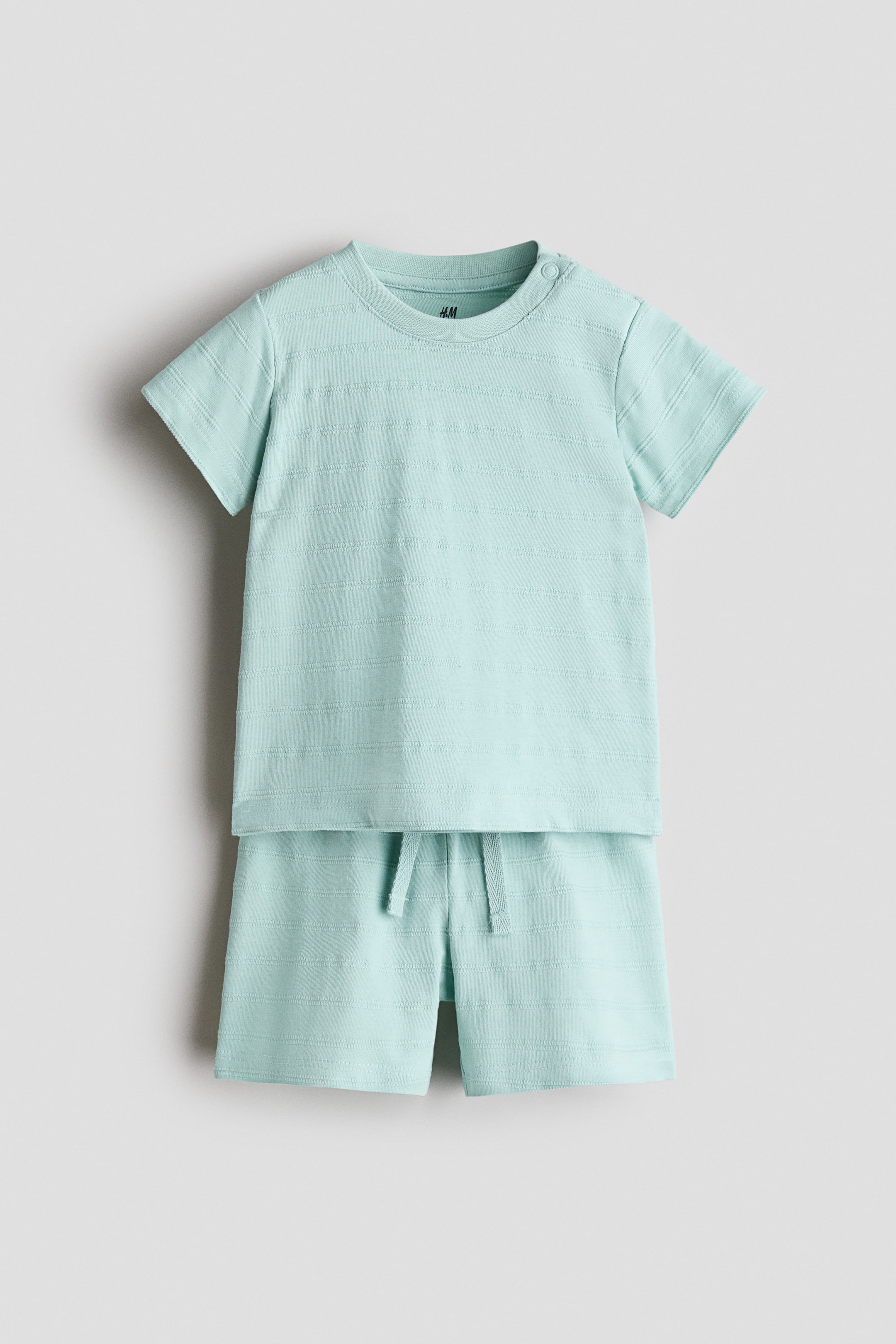 Größeres Bild ansehen: Ein hellblaues T-Shirt und Shorts-Set wird von vorne gezeigt, beide Teile weisen ein dezentes horizontales Strukturstreifenmuster auf weichem Baumwolljersey auf. Das T-Shirt verfügt über einen Rundhalsausschnitt und Druckknöpfe auf einer Schulter, während die Shorts einen elastischen Kordelzugbund zeigen.