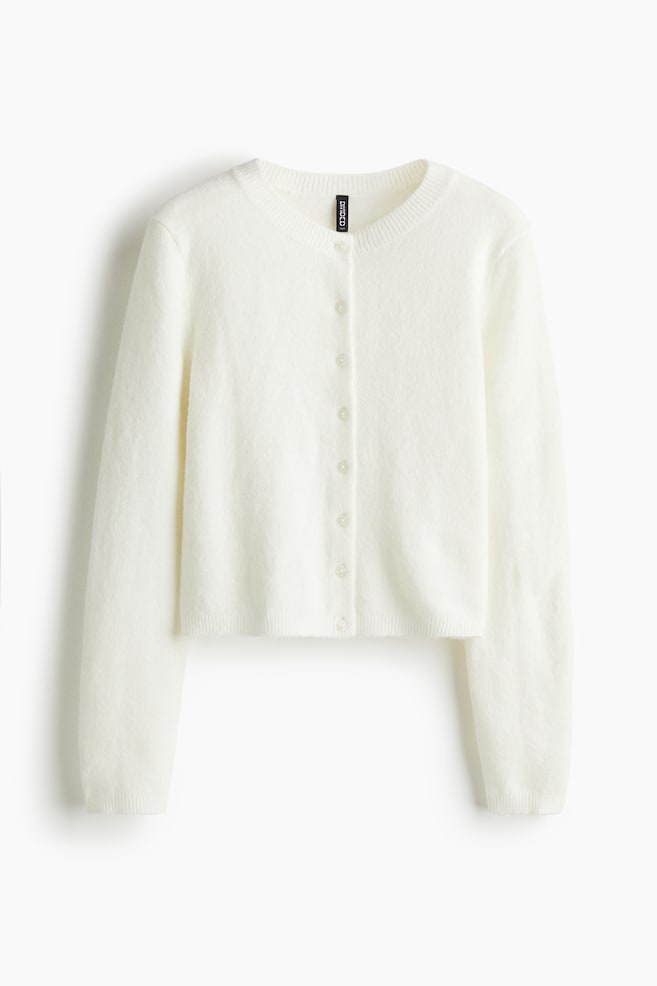 A white, short, fitted cardigan is visible from the FRONT. It is featuring a soft, fine knit, a round, ribbed neckline, and buttons running down the front. Long sleeves are completing the design, ending in ribbed cuffs and a ribbed hem. - 1