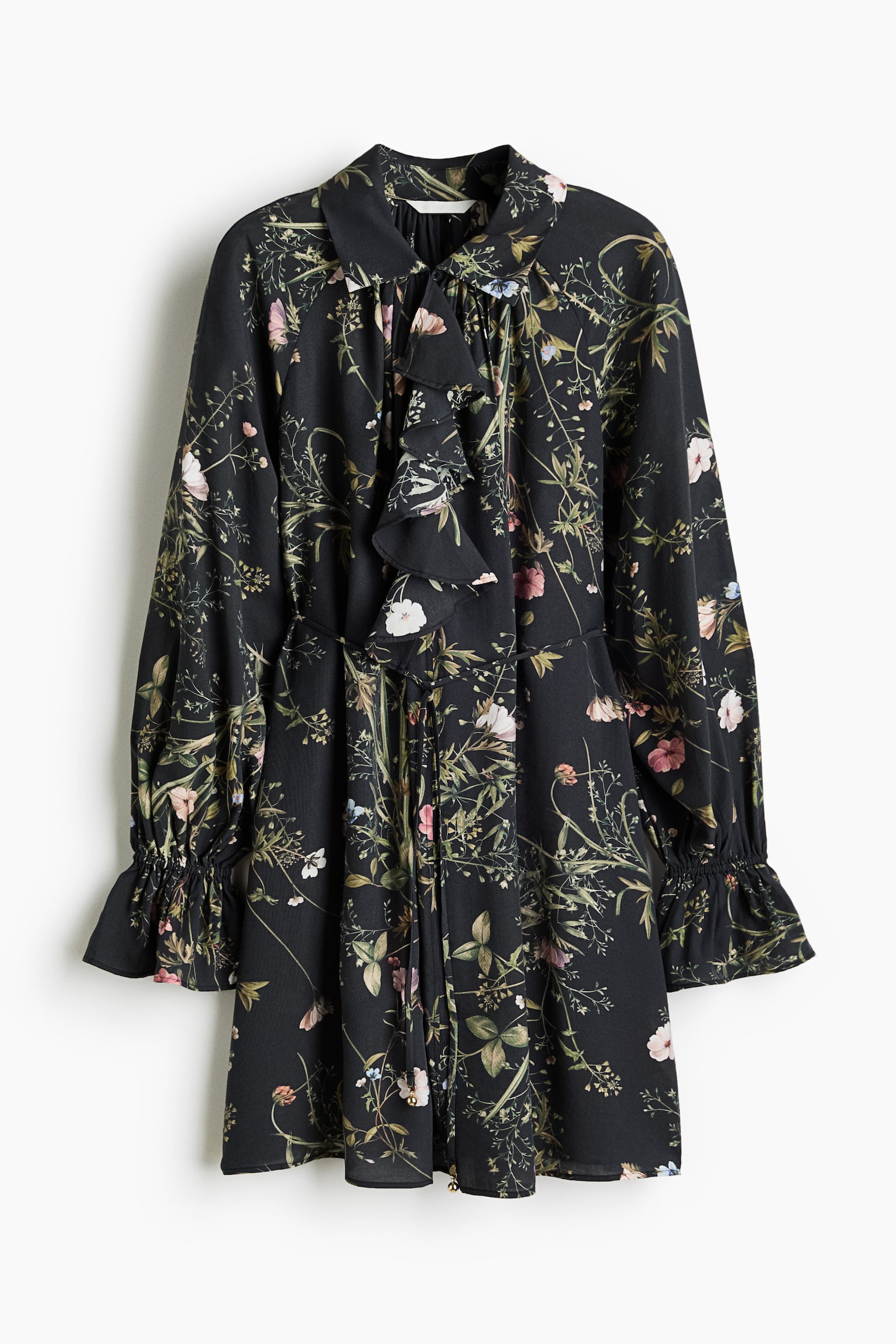 A long-sleeved mini dress, front-facing, displays an all-over botanical floral print on black fabric. The dress features a pointed collar with a ruffled front placket, a thin drawstring tie at the waist, and long balloon sleeves with gathered, ruffled cuffs.