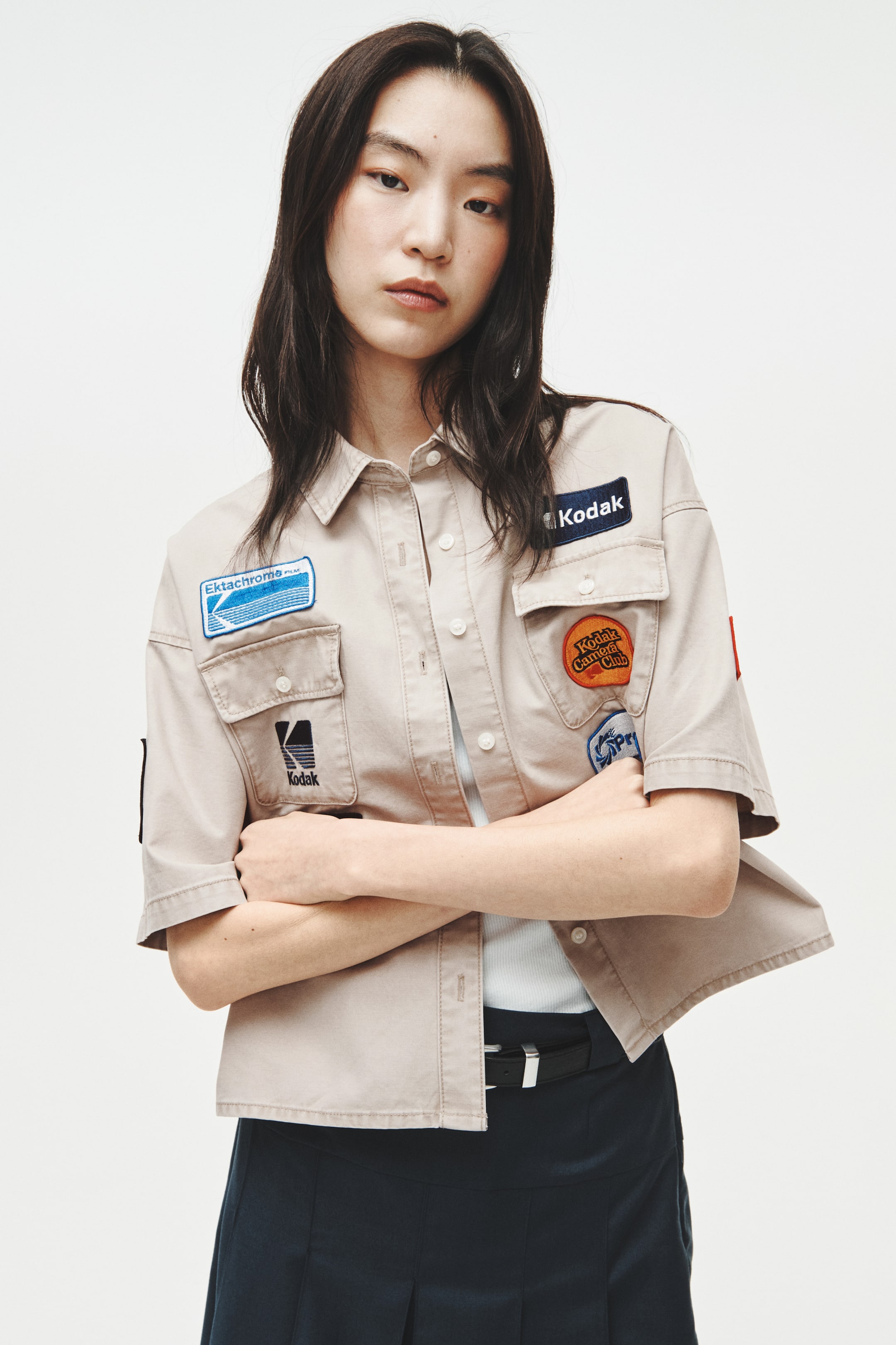 View larger image: A beige short-sleeved button-up shirt is being presented from a front angle, showing a pointed collar, a button-up front, and two flap chest pockets. It is showcasing multiple embroidered patches, including 'Kodak' and 'Ektachrome Film' on the chest and 'Kodak Camera Club' on the right sleeve, and is layered over a white ribbed top.