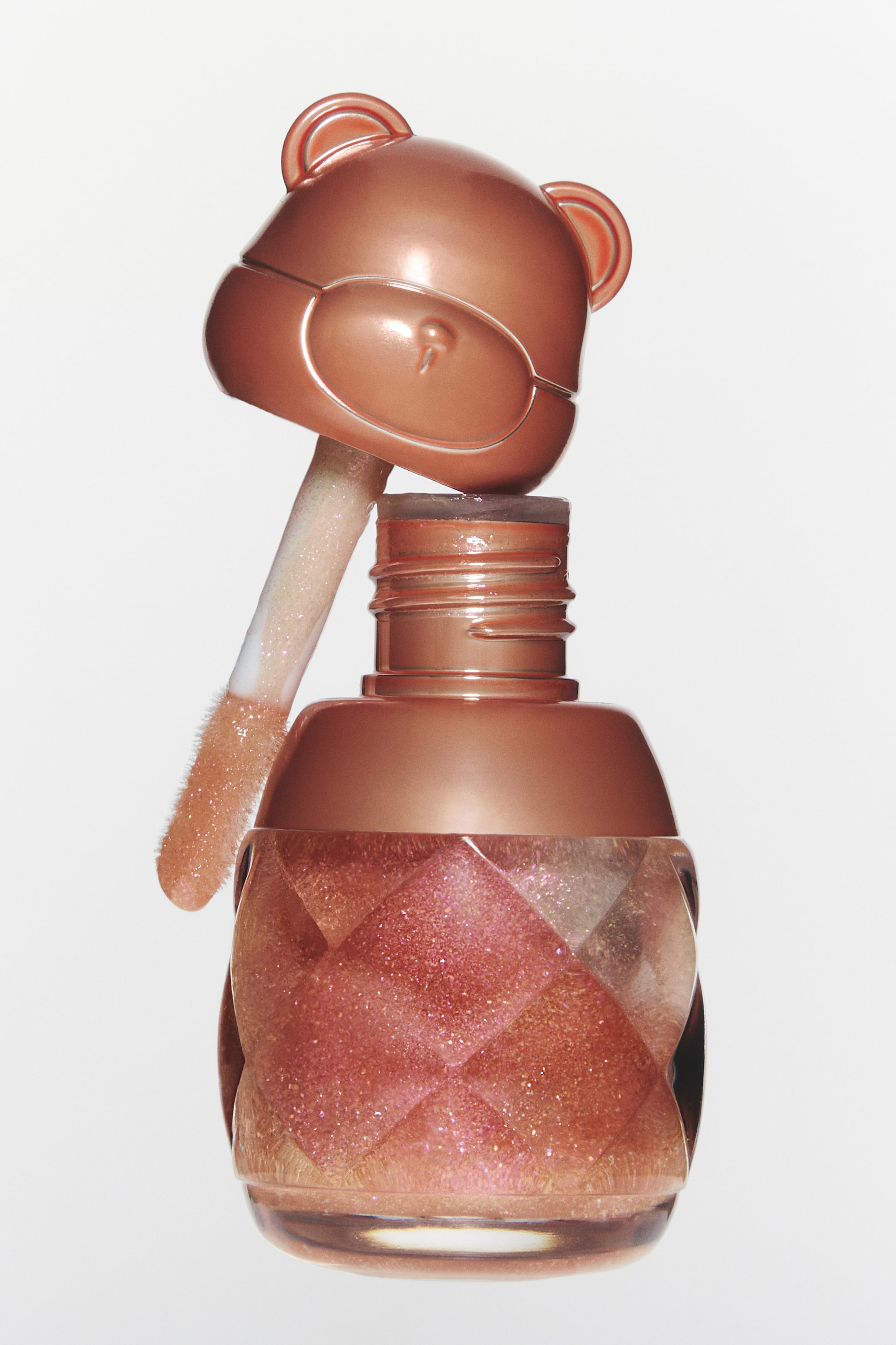 Se større billede: Et lipgloss-produkt præsenterer en roseguld bjørnehovedhætte med stiliserede briller, som hviler i en vinkel over den gevindskårne flaskehals. Flasken indeholder en skinnende ferskentonet væske og viser et quiltet mønster på sin klare krop, mens dens doe-foot applikator er belagt med den samme glitrende væske.