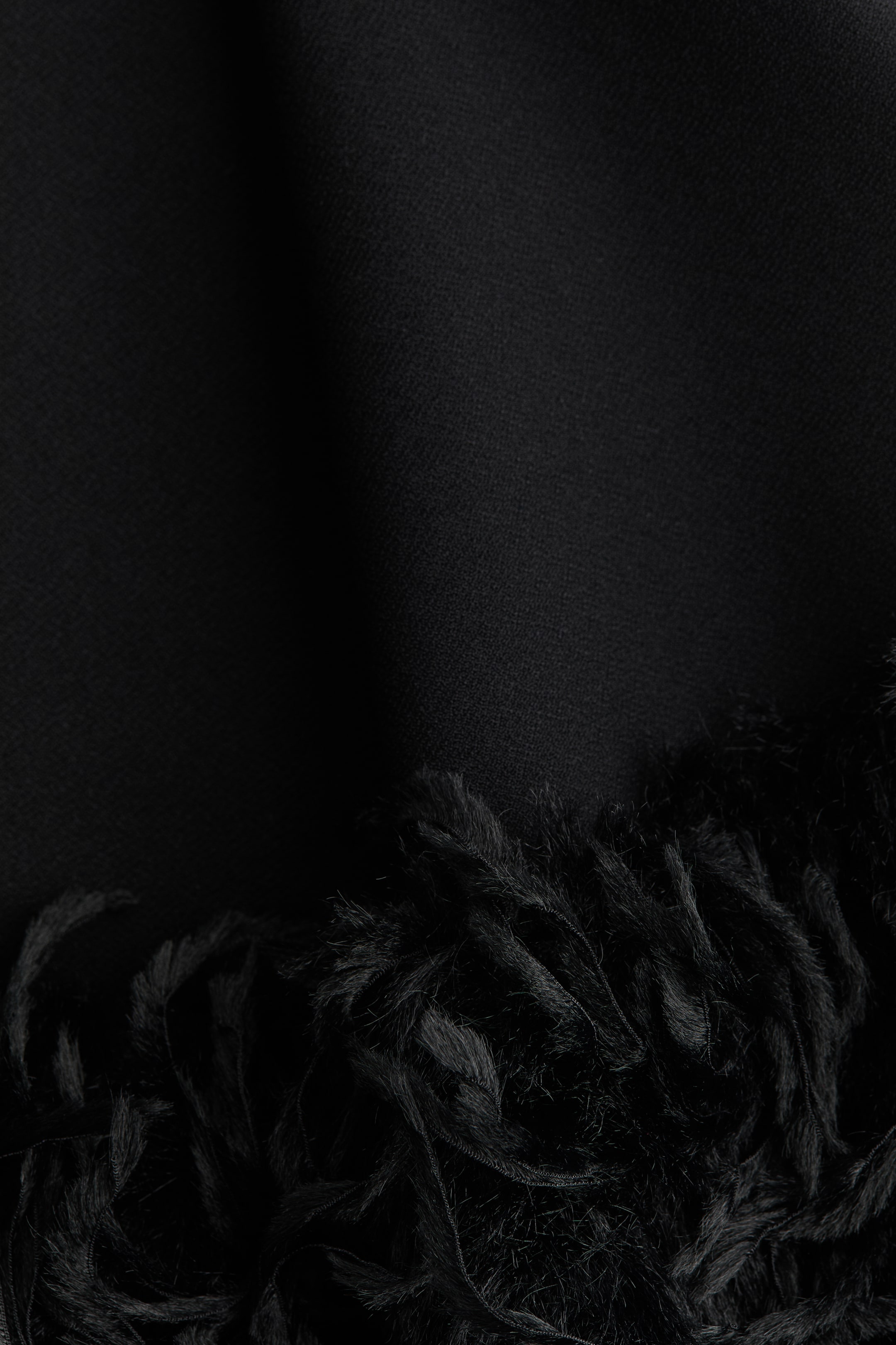 Se større billede: Et sort stof fremviser to kontrasterende teksturer; den øverste del viser en glat, fint vævet overflade, og den nederste del har et lurvet, frynset materiale med synlige tråde.