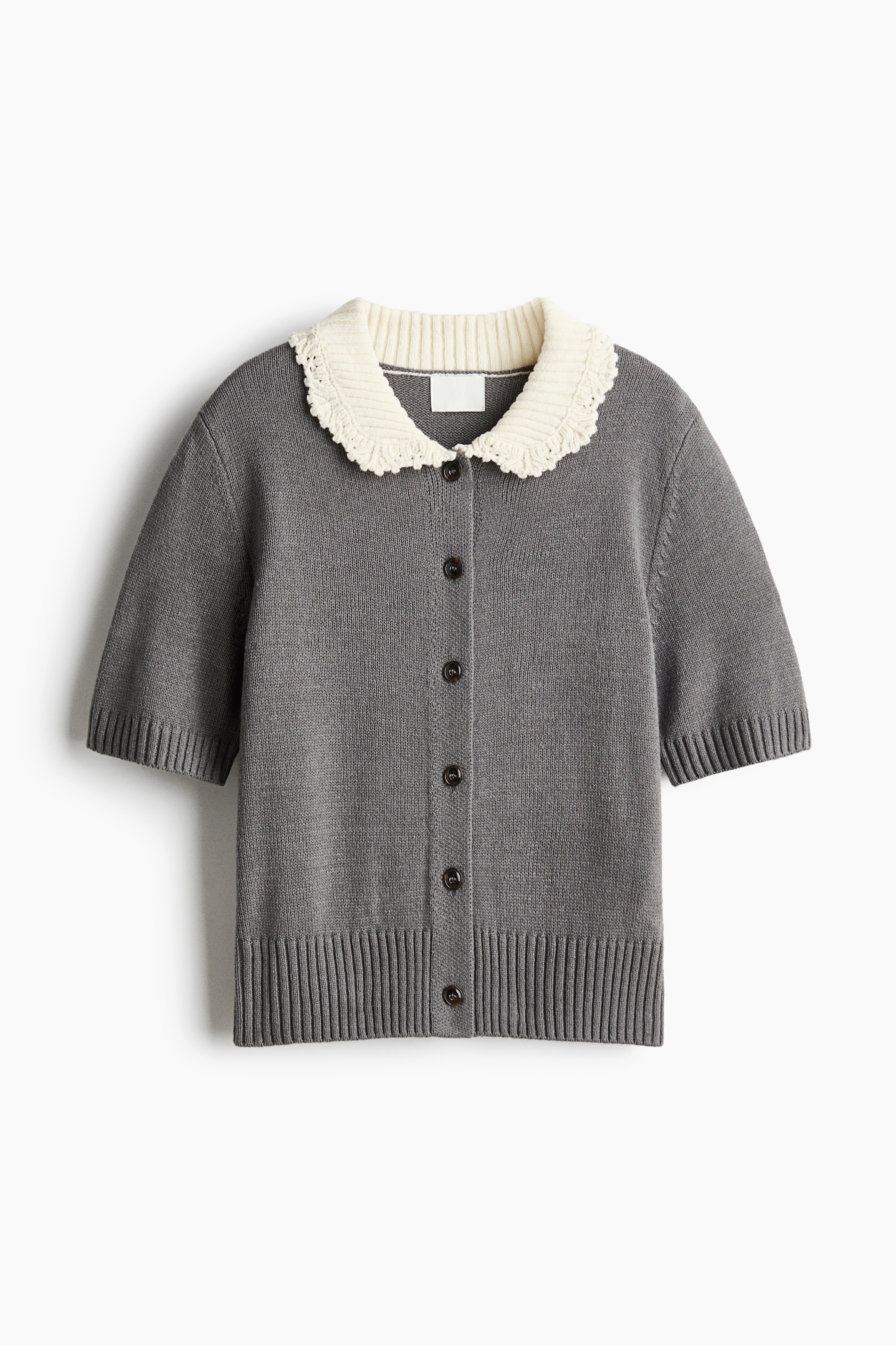 NgÆ°á»i máº«u Äang máº·c má»t chiáº¿c Ã¡o cardigan len dá»t kim tay ngáº¯n mÃ u xÃ¡m, ná»i báº­t vá»i hÃ ng cÃºc phÃ­a trÆ°á»c, cá» tay bo gÃ¢n, gáº¥u Ã¡o bo gÃ¢n báº£n rá»ng vÃ  pháº§n cá» trÃ²n mÃ u kem tÆ°Æ¡ng pháº£n vá»i viá»n dá»t kim cÃ³ káº¿t cáº¥u. Ão cardigan ÄÆ°á»£c phá»i cÃ¹ng chÃ¢n vÃ¡y midi xáº¿p ly mÃ u xanh lÃ¡ Äáº­m vÃ  bá»t da Äen cao Äáº¿n gá»i.
