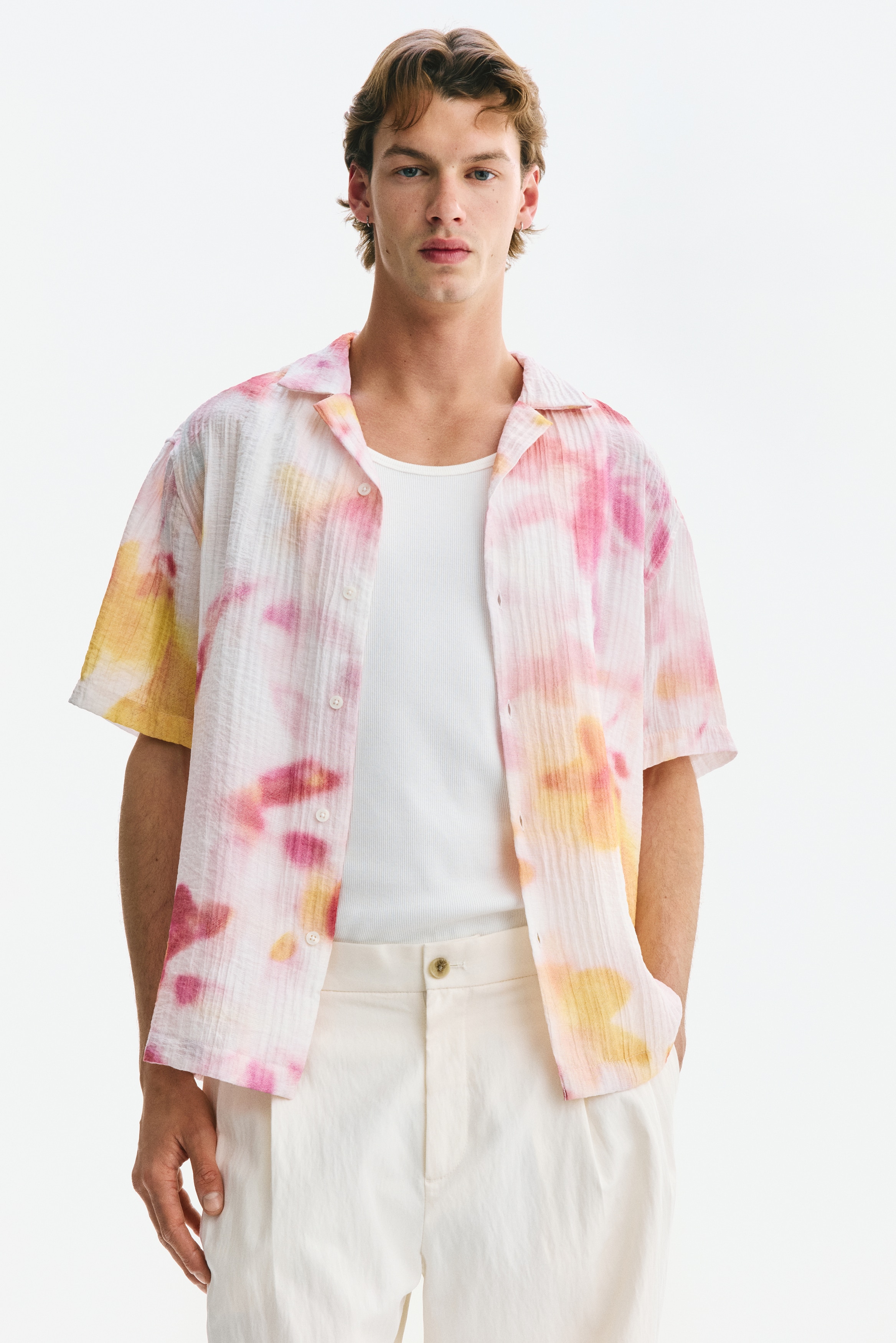 Una camisa de manga corta se presenta en vista frontal, con un tejido de algodón arrugado y patrones de tie-dye en rosa y amarillo. Incluye un cuello resort, una parte frontal francesa y un dobladillo recto con aberturas laterales.