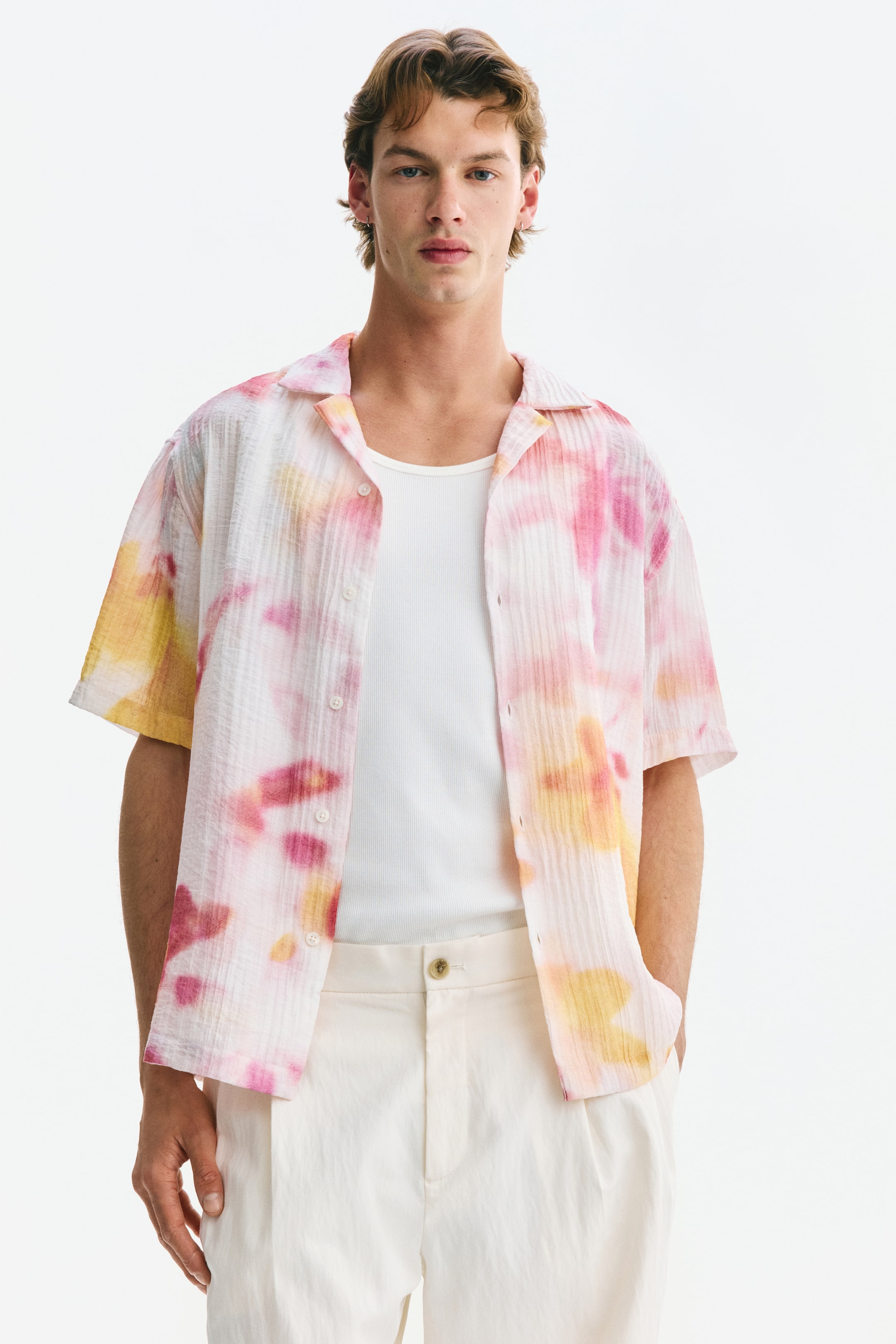 Ampliar la imagen: Una camisa de manga corta presenta un diseño de tie-dye en rosa, amarillo y blanco, con una textura arrugada y un cuello campestre. Se lleva abierta sobre una camiseta de tirantes acanalada blanca, mostrando su diseño con botones al frente.