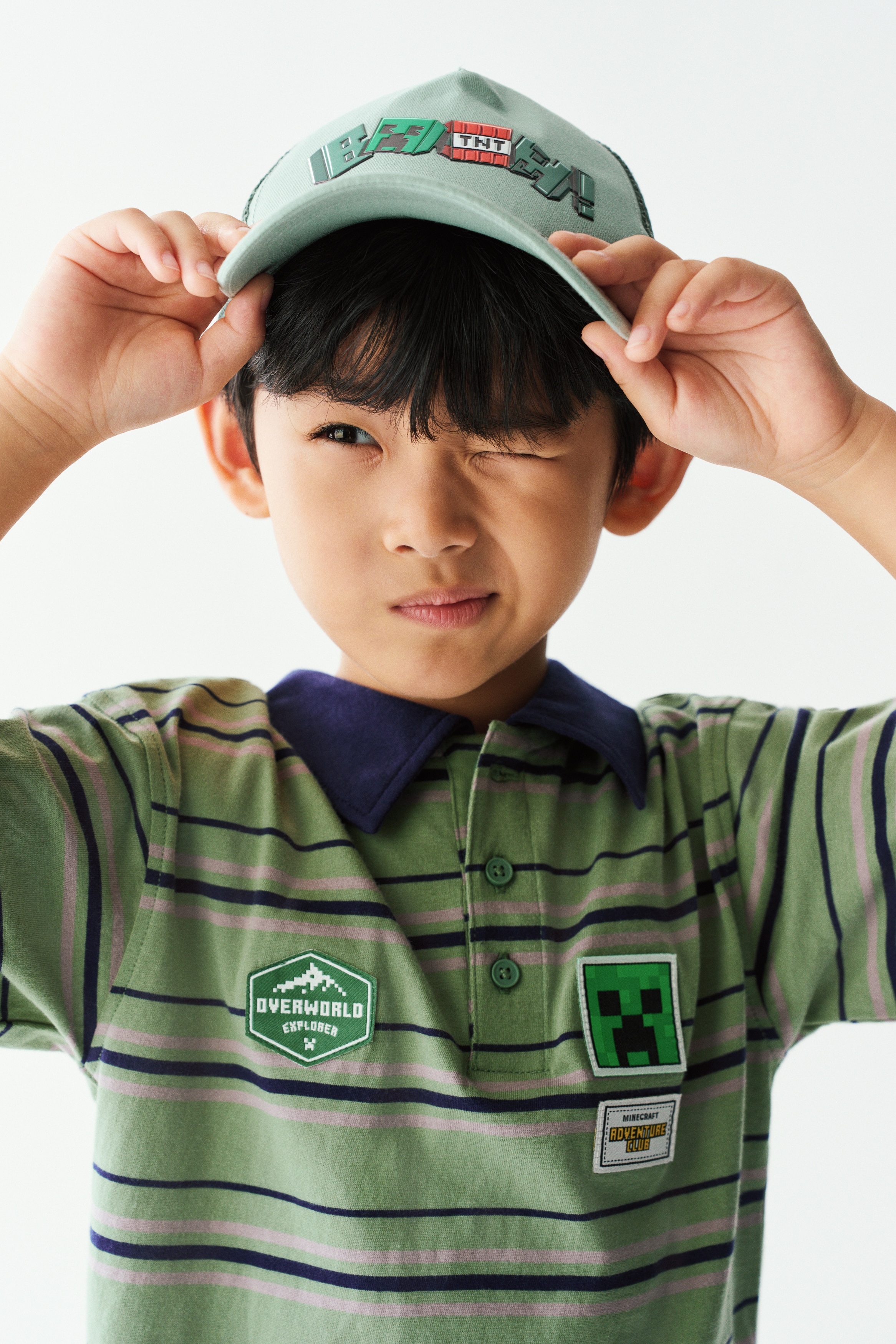 Má»t chiáº¿c Ã¡o polo dÃ¡ng há»p káº» sá»c xanh lÃ¡ vÃ  xanh navy ÄÆ°á»£c trÆ°ng bÃ y á» máº·t trÆ°á»c, ná»i báº­t vá»i cá» báº» mÃ u xanh navy tÆ°Æ¡ng pháº£n, náº¹p cÃ i ba nÃºt vÃ  vai xuÃ´i cÃ¹ng tay ngáº¯n. Pháº§n ngá»±c ÄÆ°á»£c Äiá»m xuyáº¿t cÃ¡c miáº¿ng vÃ¡ Äá» há»a chá»§ Äá» Minecraft, bao gá»m khuÃ´n máº·t Creeper mÃ u xanh lÃ¡ vÃ  huy hiá»u âOverworld Explorerâ.