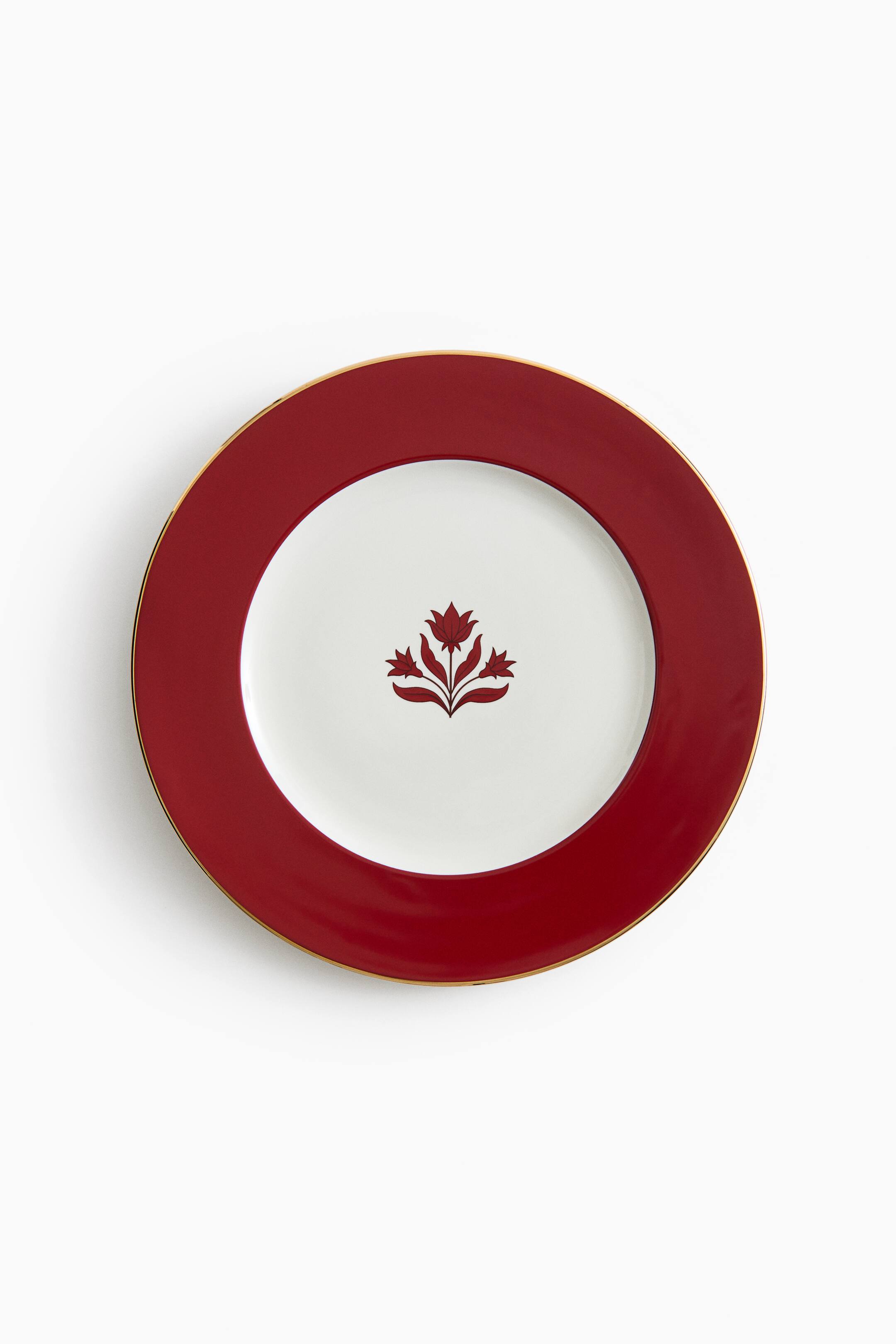 View larger image: A round ceramic plate is lying flat in a top-down view, featuring a wide, deep red rim with a thin gold trim. The white centre of the plate displays a stylised dark red floral motif of three blossoms.