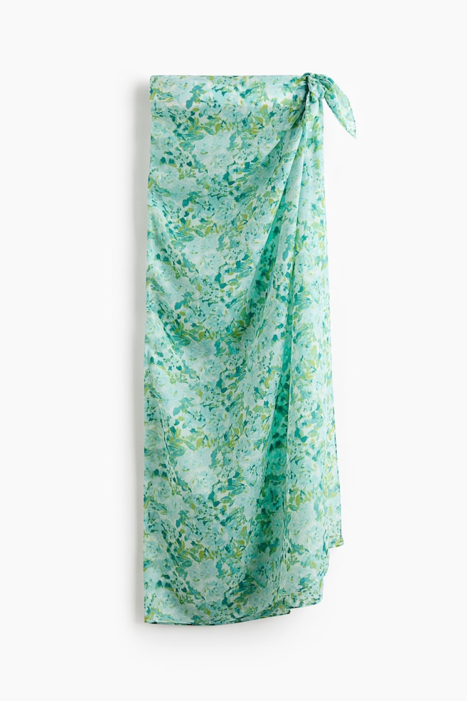 A long sarong in airy voile is displaying a green and blue mottled print, and is being tied at the front, forming a skirt. - 1