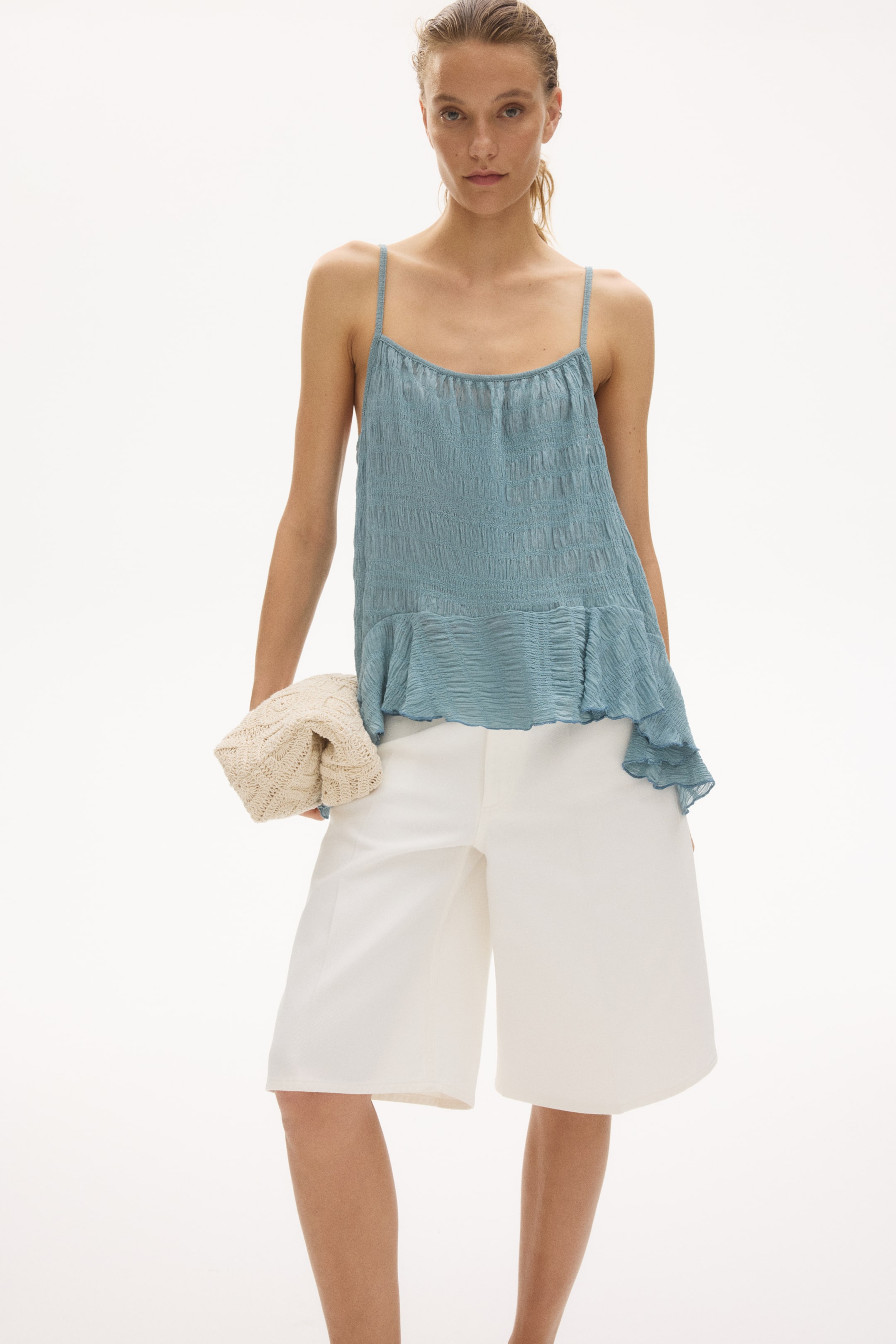 Ampliar la imagen: Se presenta una blusa de tirantes azul desde el frente, con finos tirantes tipo espagueti, una textura arrugada y un dobladillo asimétrico con volantes. Se combina con shorts blancos de pierna ancha que se extienden justo por encima de la rodilla.