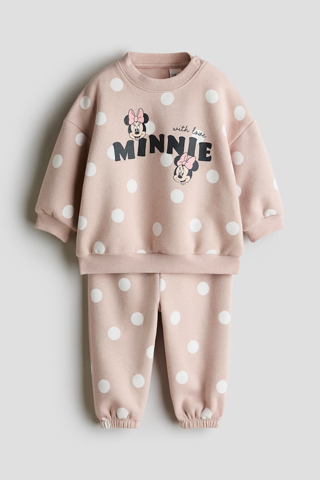 Et 2-delt sæt i printet sweatstof i lyserosa med hvide prikker er vist forfra og består af en langærmet sweatshirt og matchende sweatpants. Sweatshirten har et Minnie Mouse-tryk med teksten "MINNIE" på brystet samt ribbet rund hals, ribbede manchetter og ribkant forneden, mens sweatpantsene har elastiske opsmøgede benafslutninger. - 1