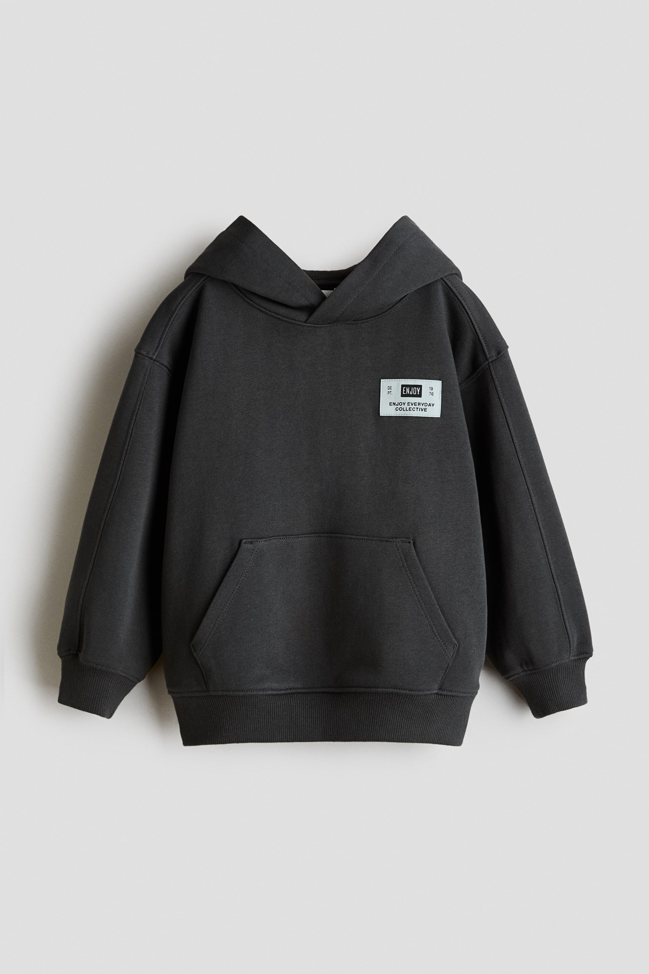View larger image: A dark grey hoodie is shown from the front, featuring a prominent hood, a large kangaroo pocket, and ribbed cuffs and hem. A light-coloured rectangular label with the text "ENJOY" is affixed to the left chest.