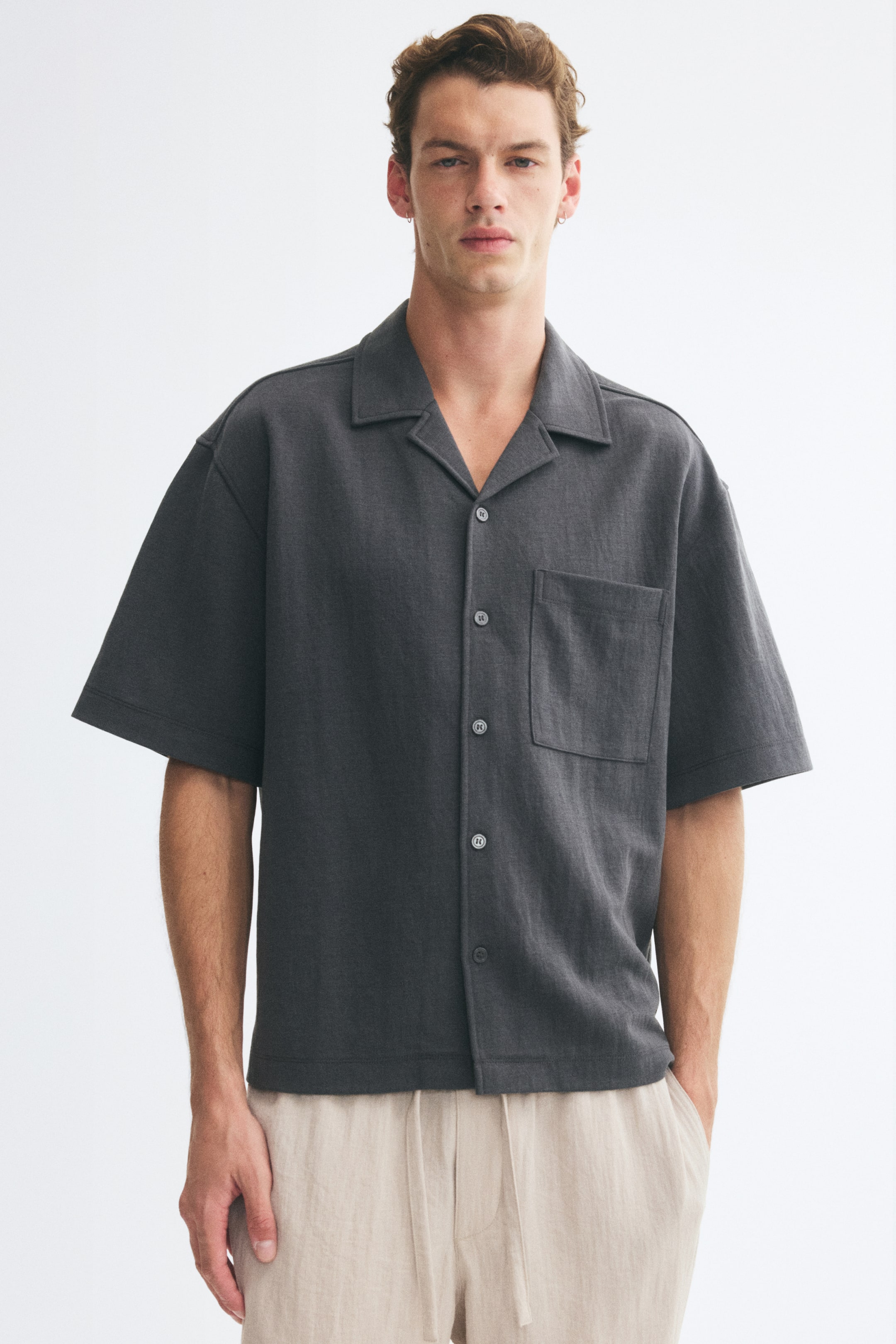 View larger image: The model is wearing a dark grey short-sleeve shirt in a relaxed fit, featuring a resort collar, button front, and a single patch chest pocket. The shirt is styled with light-colored drawstring trousers.