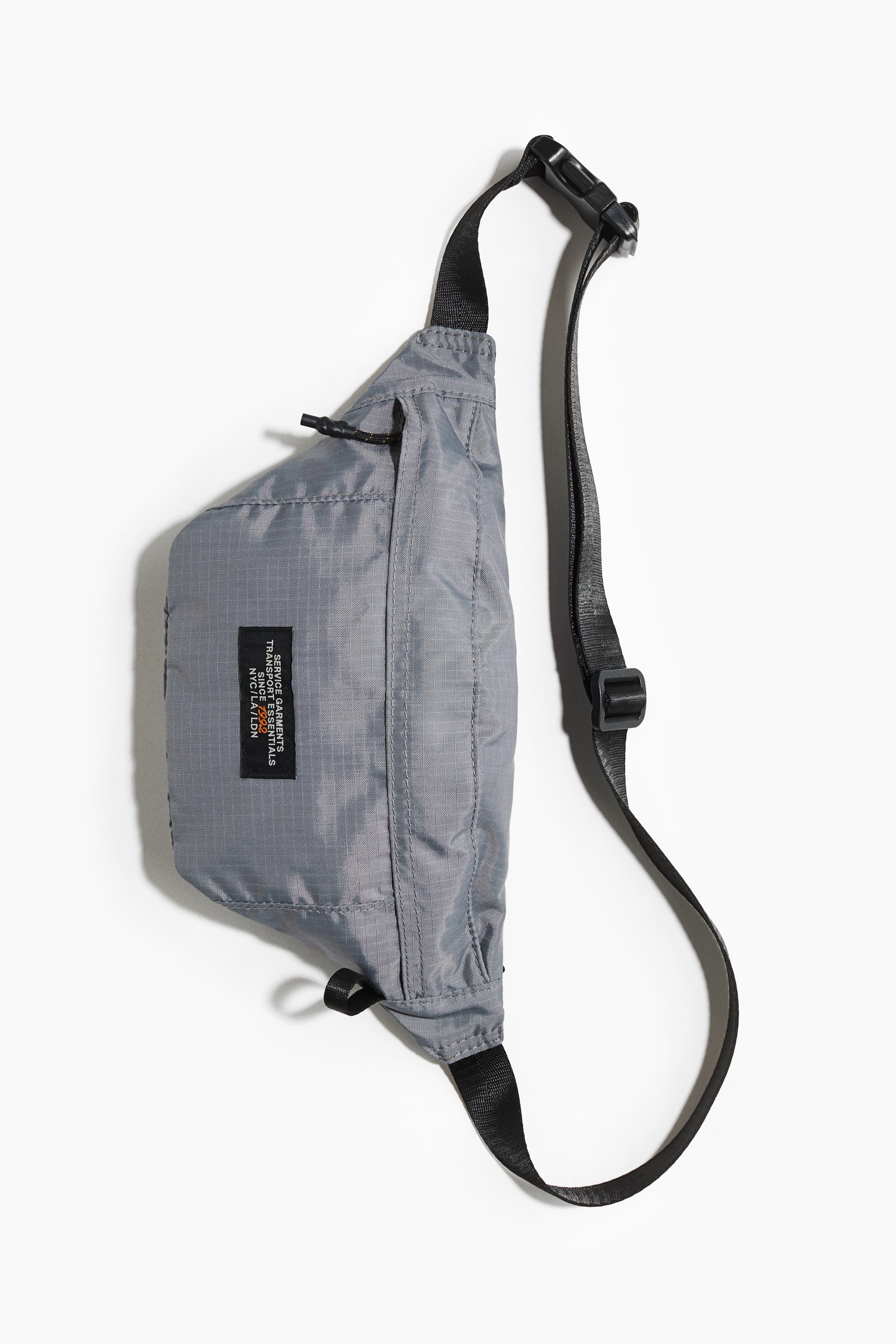Ver imagen más grande: Un bolso de bandolera gris presenta su PARTE FRONTAL, hecho de tela ripstop y con una correa ajustable negra con hebilla de plástico. Se aprecia una etiqueta de tela negra en su superficie, junto con un tirador de cremallera de cordón negro en la parte superior.