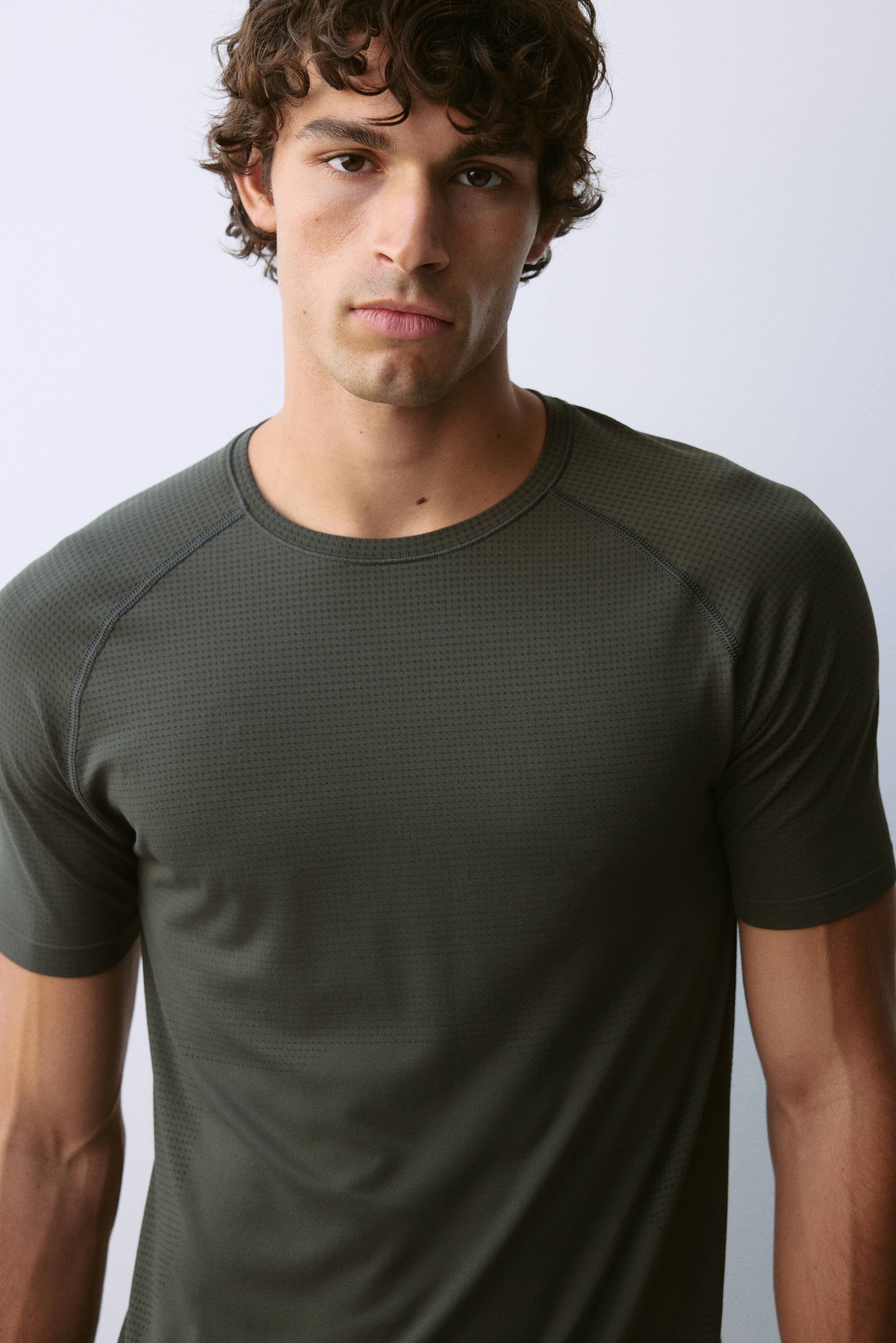 View larger image: A dark olive green athletic t-shirt is being worn by the model, featuring a crew neck and short raglan sleeves. The garment is constructed from a fine, micro-perforated textured fabric with flatlock seams.