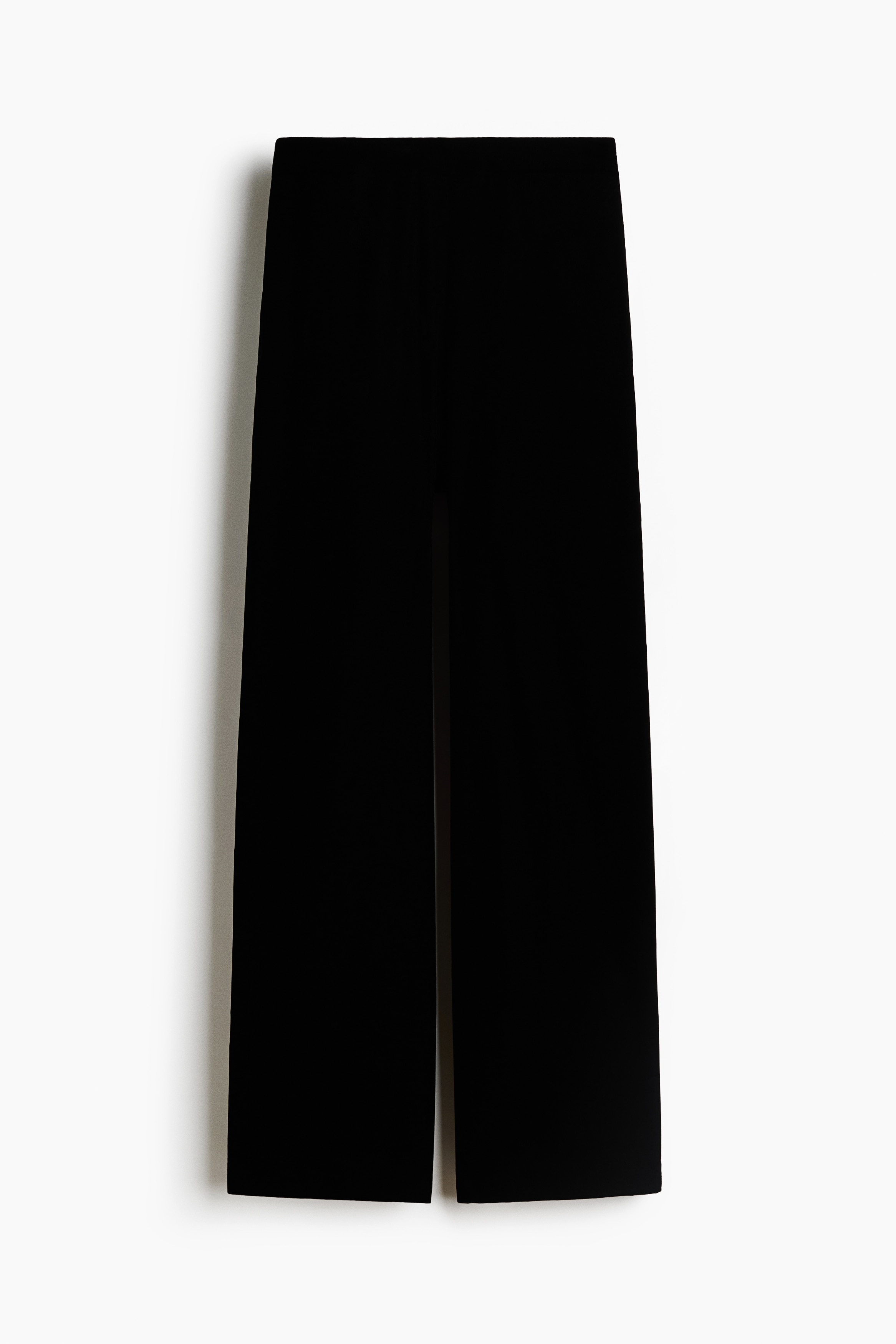 Un pantalon en velours noir à la silhouette longue et large est porté par le mannequin, qui se tient debout, les mains dans les poches latérales. Le pantalon tombe entièrement sur les pieds, laissant apparaître en partie des chaussures noires à bout pointu, et est assorti à une veste en velours noir coordonnée.