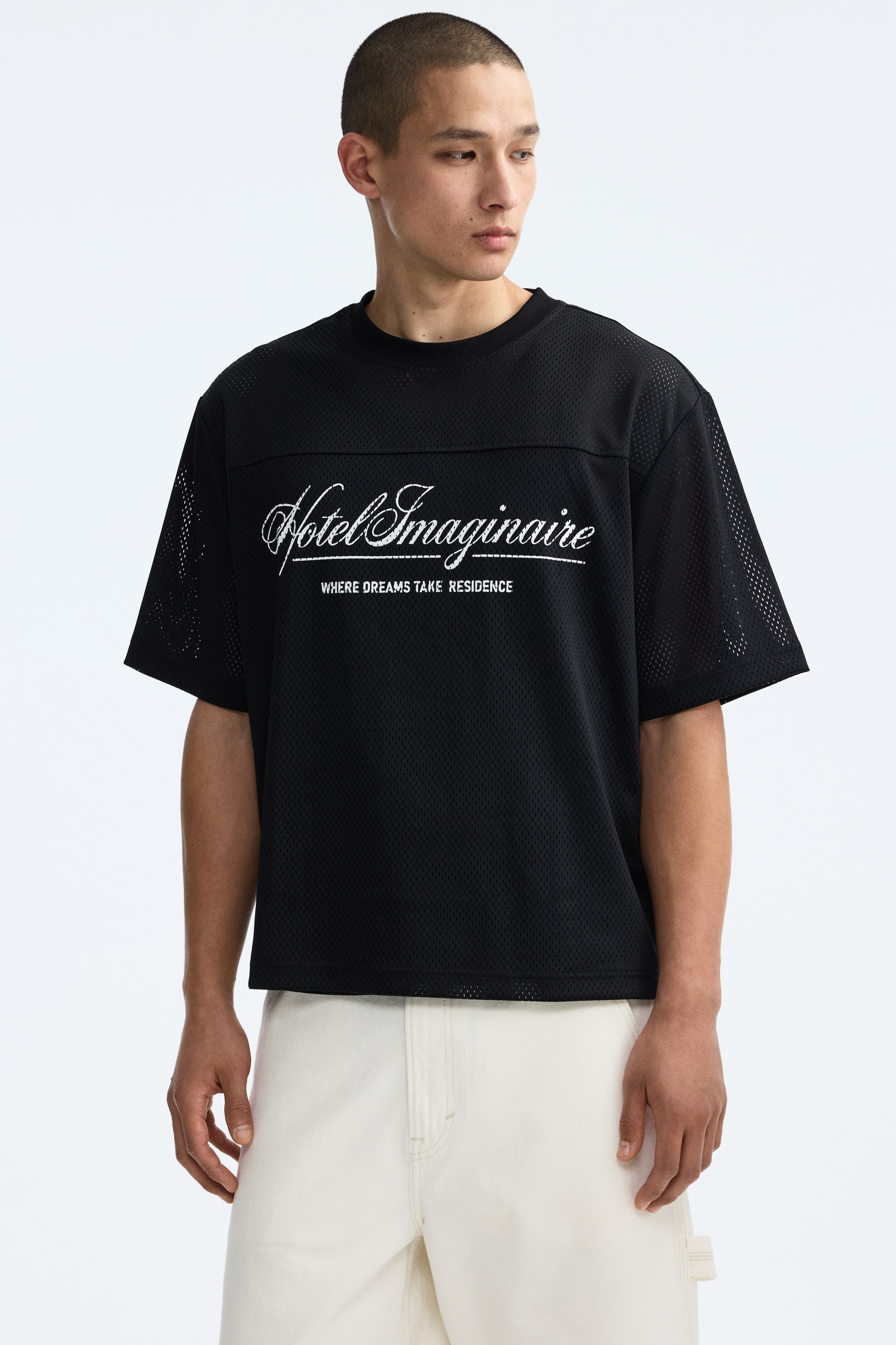 View larger image: The model is wearing a black mesh T-shirt with a white "Hotel Imaginaire" print across the chest. It features a round neckline, dropped shoulders, a front yoke, and a straight-cut hem, providing an oversized, baggy fit.
