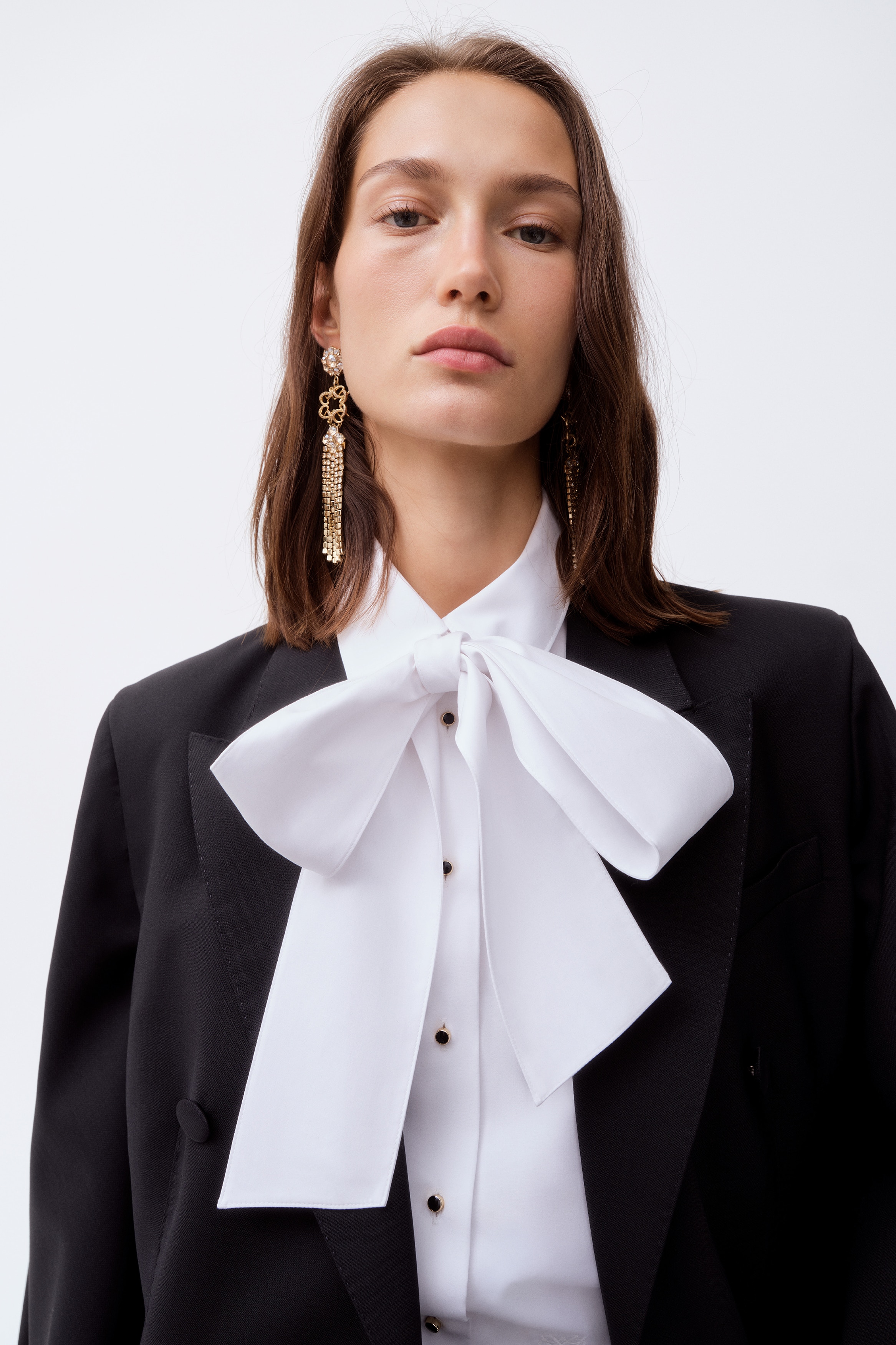 The Bow Collar Tuxedo Shirt is shown in a front-facing view, featuring a large, structured bow tied at the high collar. The shirt showcases a white bib-front panel accented with small dark stud buttons, long sleeves with wide cuffs, and a subtly gathered waistline that defines the silhouette.