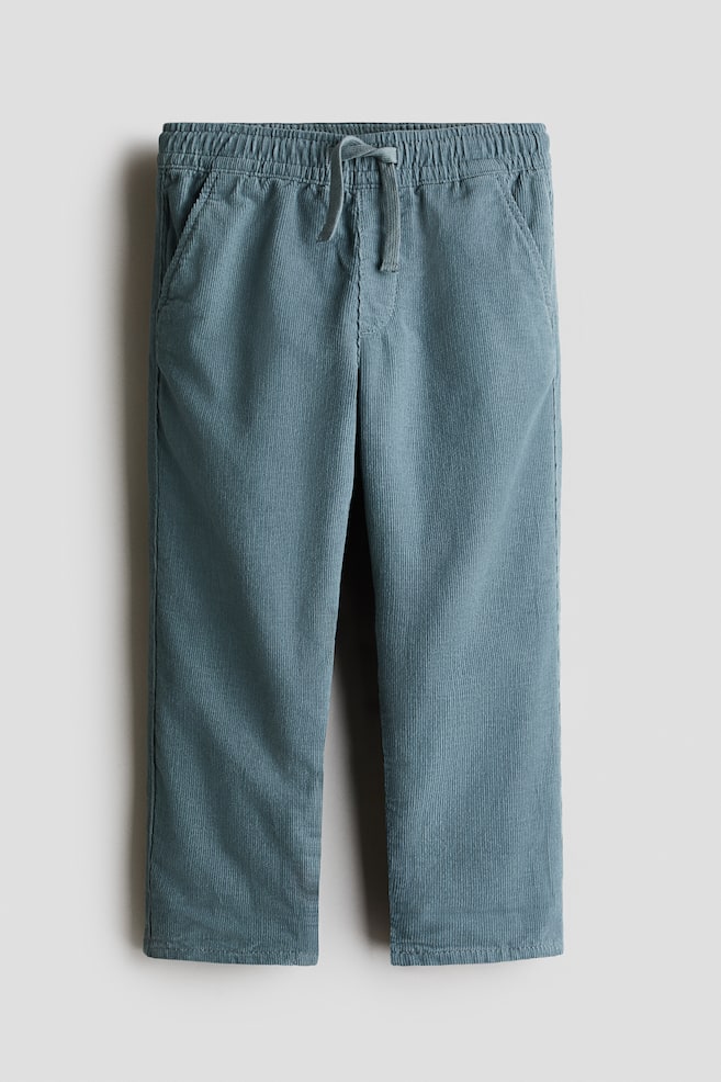 A front view of a pair of dusty blue corduroy pants featuring a textured, ribbed fabric and a relaxed fit with gently tapered legs. The design includes an elasticized waistband with a matching drawstring, a mock fly, and side pockets. - 1