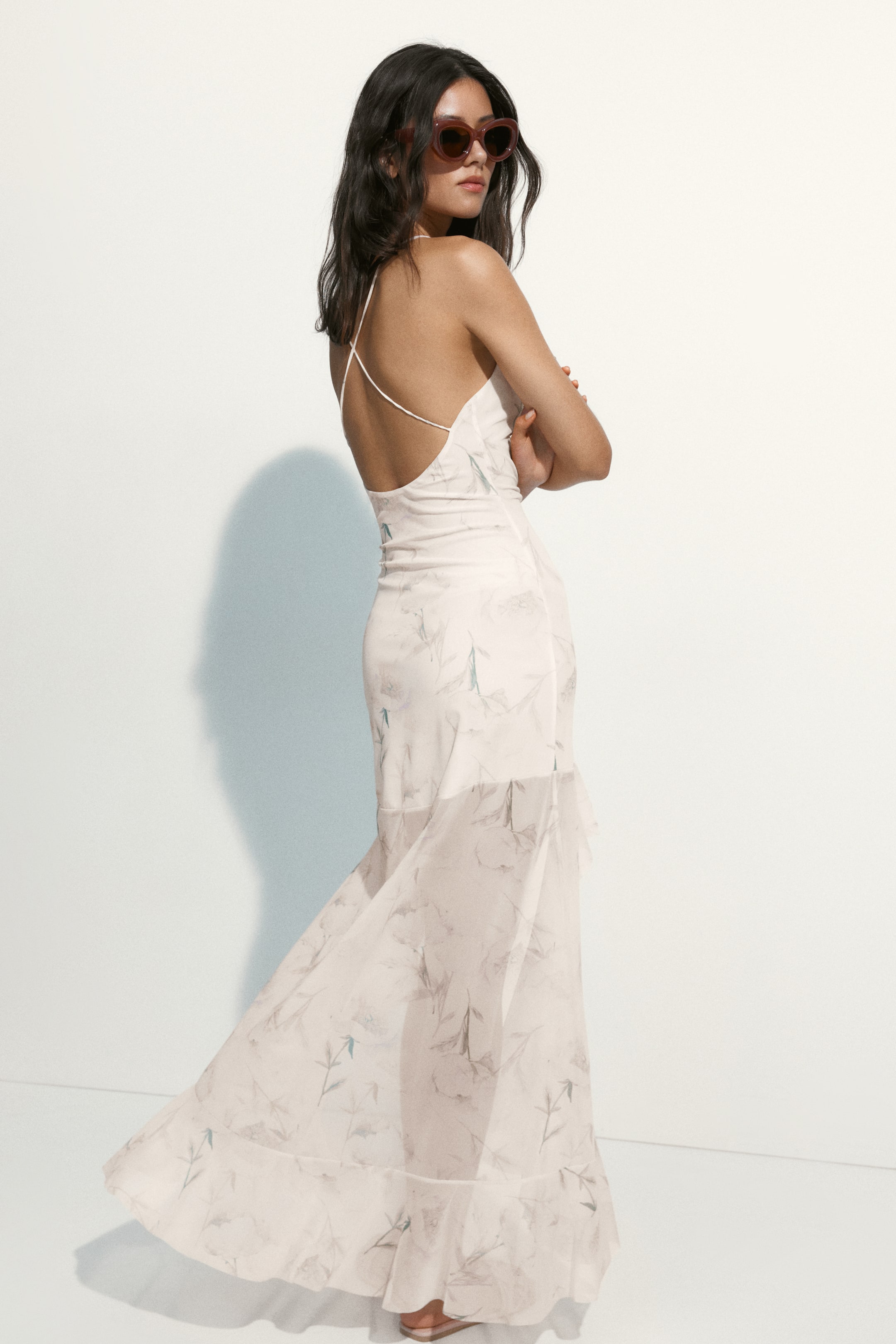 View larger image: A light-colored maxi dress showcasing a subtle gray and blue botanical pattern, thin crisscross spaghetti straps, and a low open back. The dress fits through the bodice and waist, then flows into a semi-sheer skirt with a flounce detail at the hem.