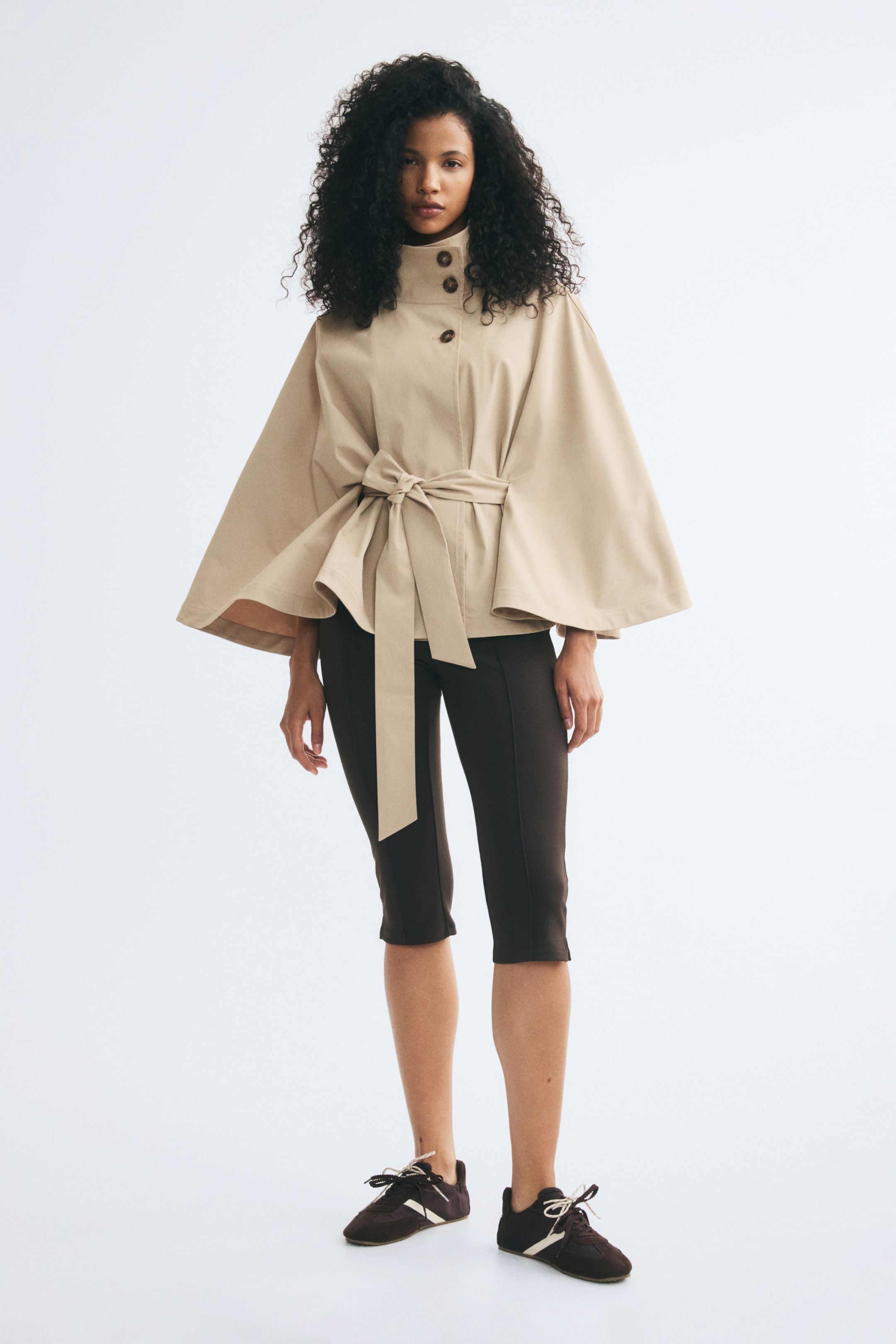 Agrandir l'image: Une veste style cape en sergé beige est portée par le mannequin, avec un col cheminée haut à détails boutonnés et de larges manches évasées. La veste est cintrée par une large ceinture assortie nouée en nœud à la taille, et associée à un pantalon cropped foncé.