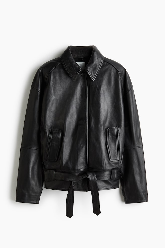 Una giacca in pelle nera Ã¨ mostrata frontalmente, mettendo in risalto una silhouette rilassata con spalle scese e un ampio colletto a punta. Presenta una chiusura frontale con bottoni nascosti, tasche verticali a filetto con patte decorative e una cintura abbinata in pelle annodata in vita. - 1