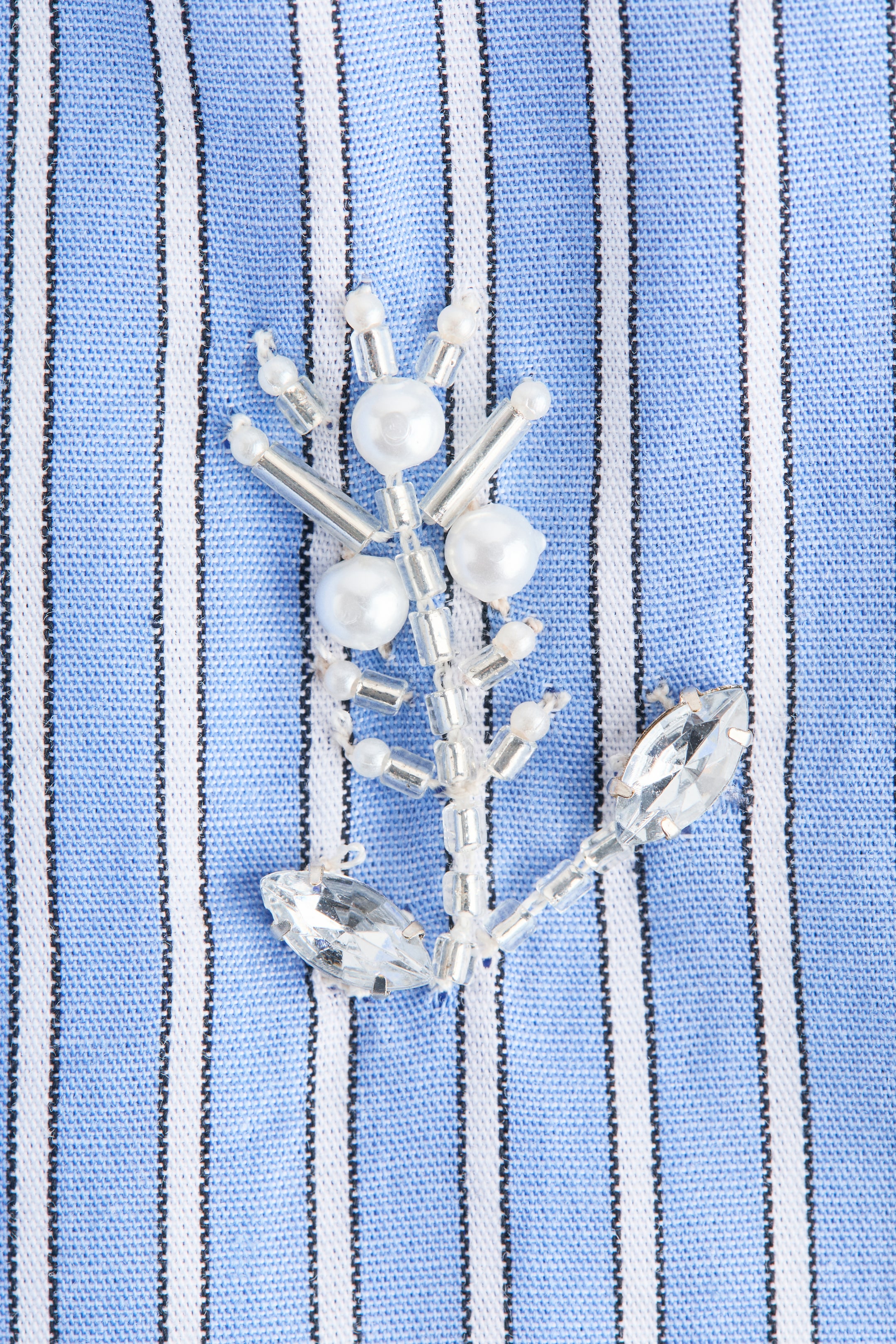 Voir une image plus grande: Un tissu à rayures bleu et blanc présente un ornement perlé. Le tissu arbore des rayures alternées bleu et blanc, avec de fines lignes noires bordant les rayures blanches. L'ornement forme un motif complexe et ramifié, orné de perles rondes blanches semblables à des perles, de perles cylindriques de couleur argentée, de minuscules perles de rocaille transparentes, et de cristaux transparents taillés en forme de marquise à ses extrémités inférieures.
