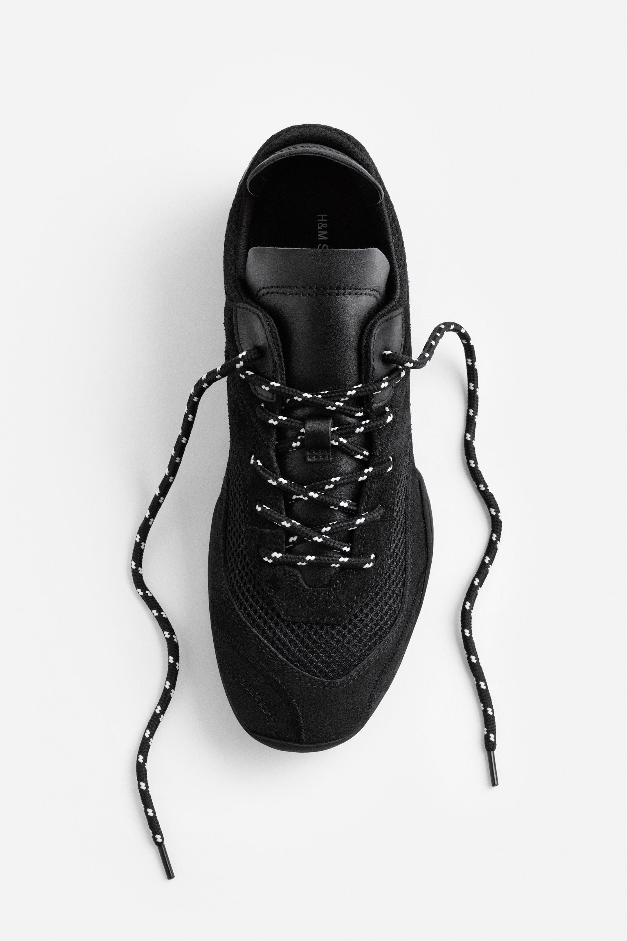 View larger image: "A black sneaker is facing forward, seen from above, featuring mesh panels on the toe box and sides, with suede overlays. Black and white speckled laces are criss-crossing over a smooth tongue, with their ends resting loosely."
