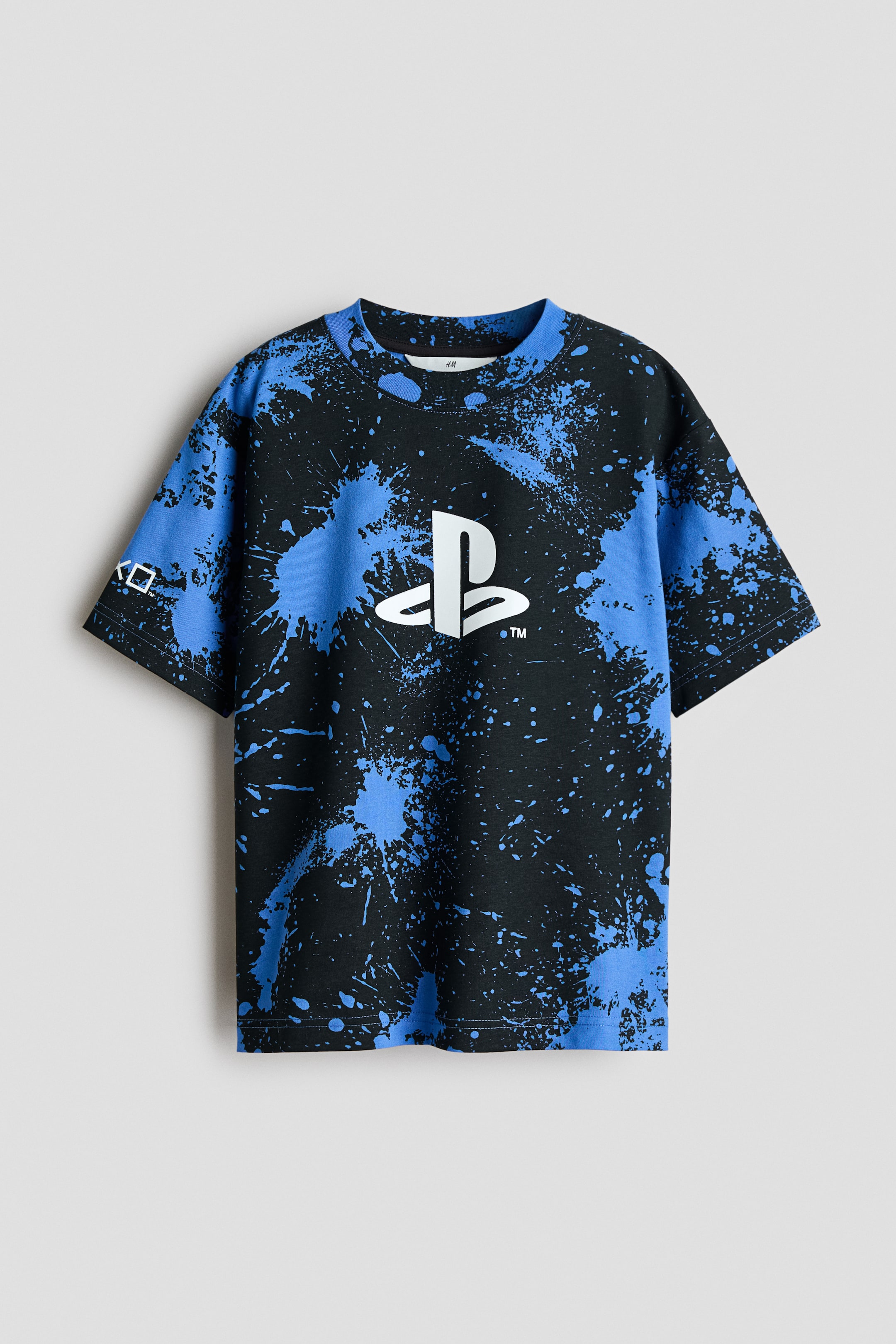 Voir une image plus grande: Vue de face d’un t-shirt en jersey de coton présentant un motif d’éclaboussures de peinture noire et bleue avec un logo PlayStation blanc imprimé au centre de la poitrine. Le t-shirt est doté d’un col rond avec bord-côte et d’épaules tombantes.