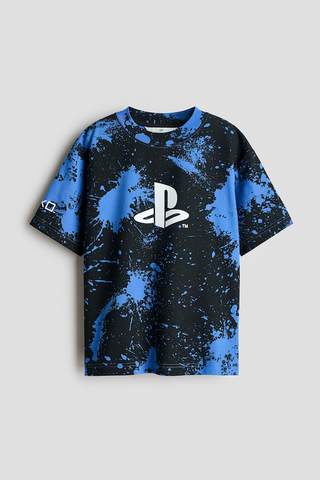 A front view of a cotton jersey t-shirt is displaying a black and blue paint-splatter design with a white PlayStation logo printed on the center chest. The tee is featuring a round, rib-trimmed neckline and dropped shoulders. - 1