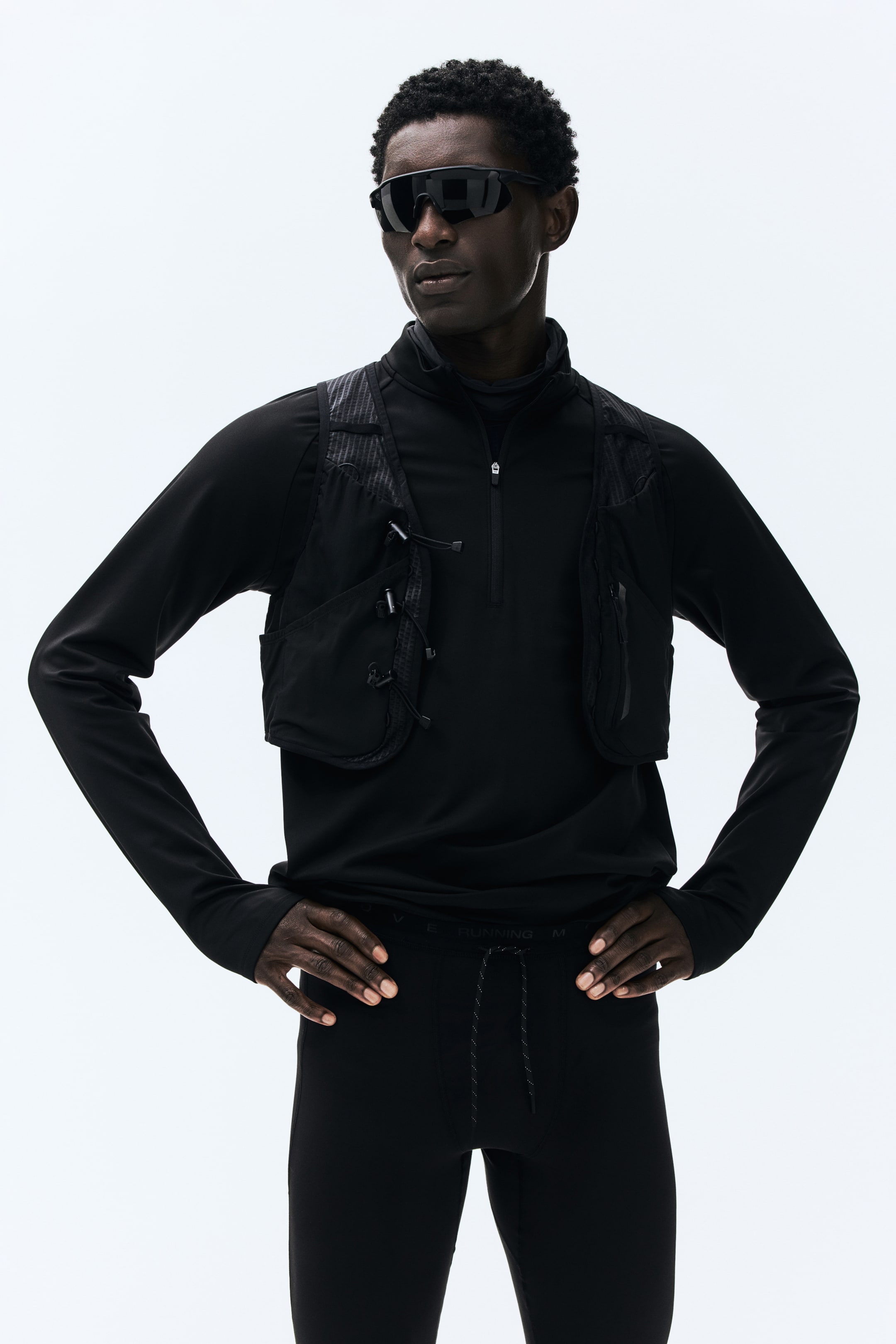 Ampliar la imagen: Un top deportivo negro de manga larga con cuello alto quarter-zip está colocado en capas debajo de un chaleco utilitario negro. El chaleco presenta múltiples bolsillos con detalles de cordón y paneles texturizados, y unos leggings negros ajustados muestran una cintura con cordón con el texto visible 'running'.