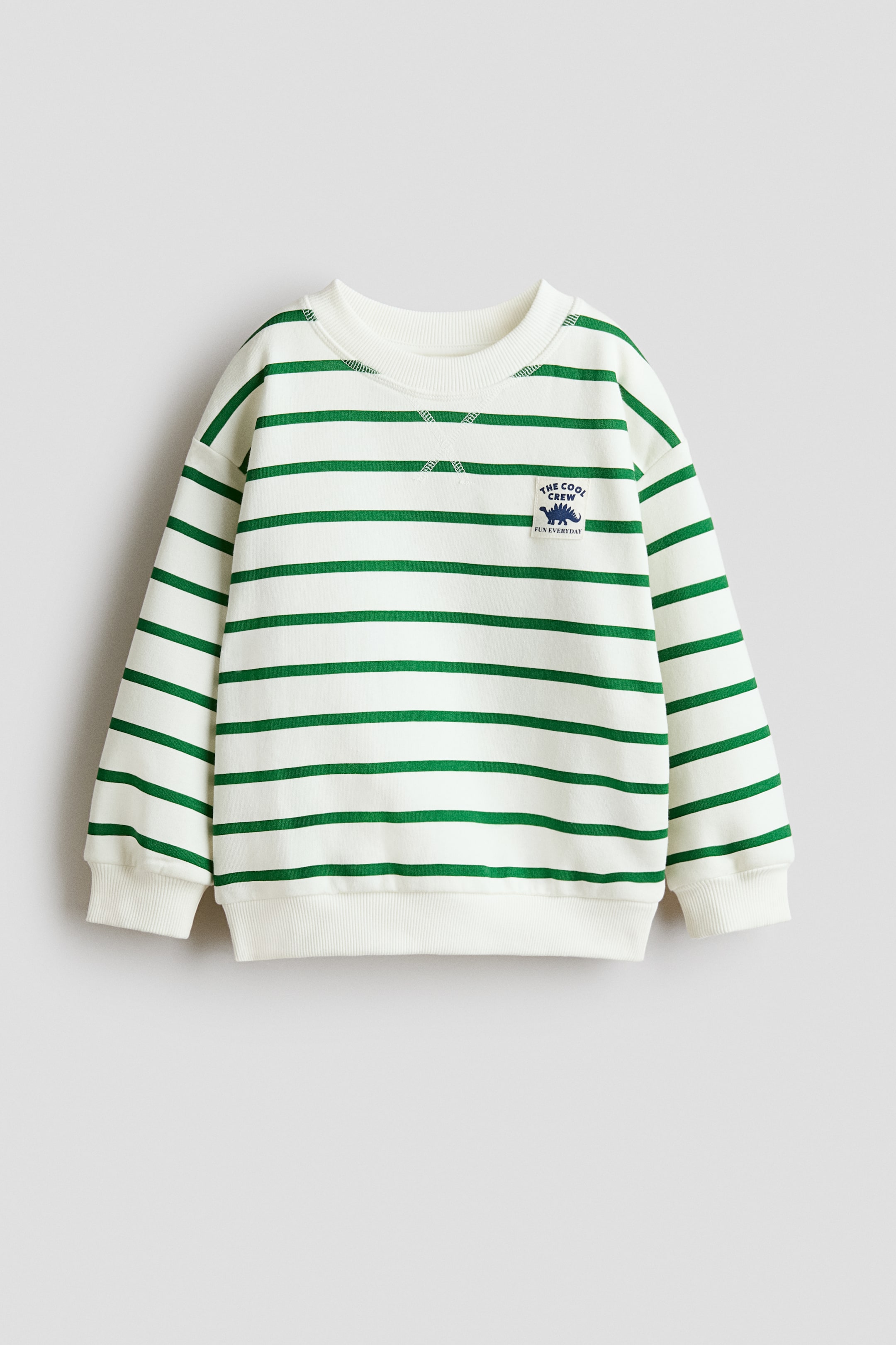 View larger image: A cream sweatshirt featuring horizontal green stripes, a round rib-trimmed neckline with crisscross stitching, dropped shoulders, long sleeves, and ribbed cuffs and hem. A small embroidered patch of a blue dinosaur with the text "THE COOL CREW FUN EVERYDAY" is visible on the left chest.
