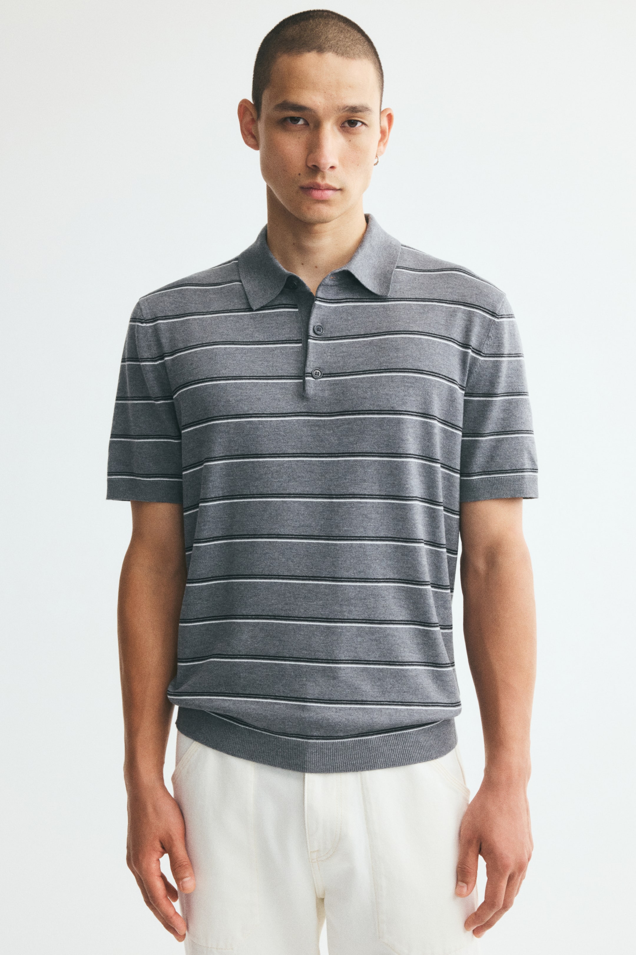 View larger image: The model is wearing a gray knit polo shirt with thin horizontal black and white stripes. The shirt features a classic collar, a button placket, short sleeves, and a ribbed hem, and is styled with white trousers.
