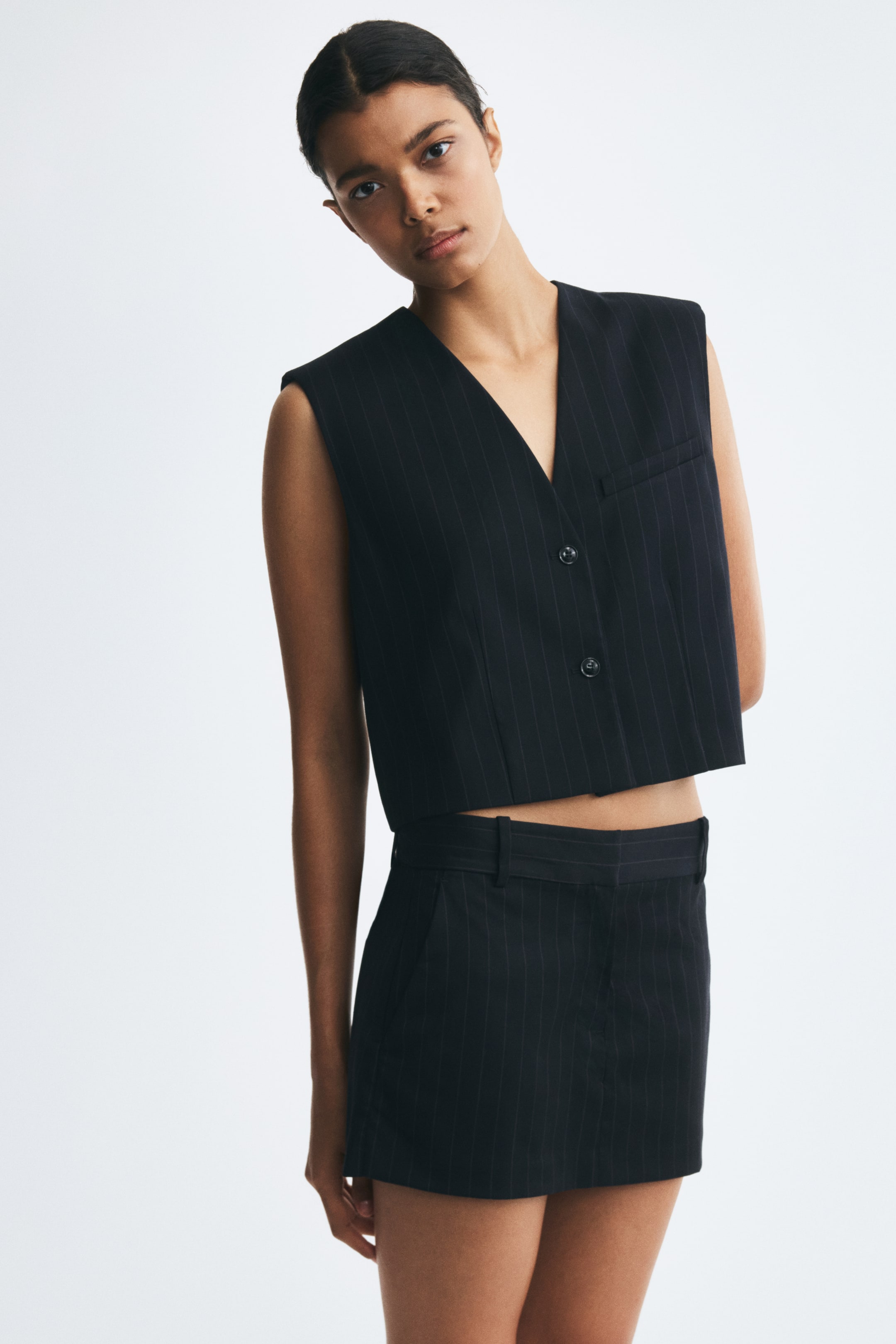 View larger image: The model is wearing a cropped black pinstripe vest featuring a deep V-neckline, structured shoulders, and a front button closure. The sleeveless vest includes a single welt chest pocket and is paired with a matching pinstripe mini skirt.